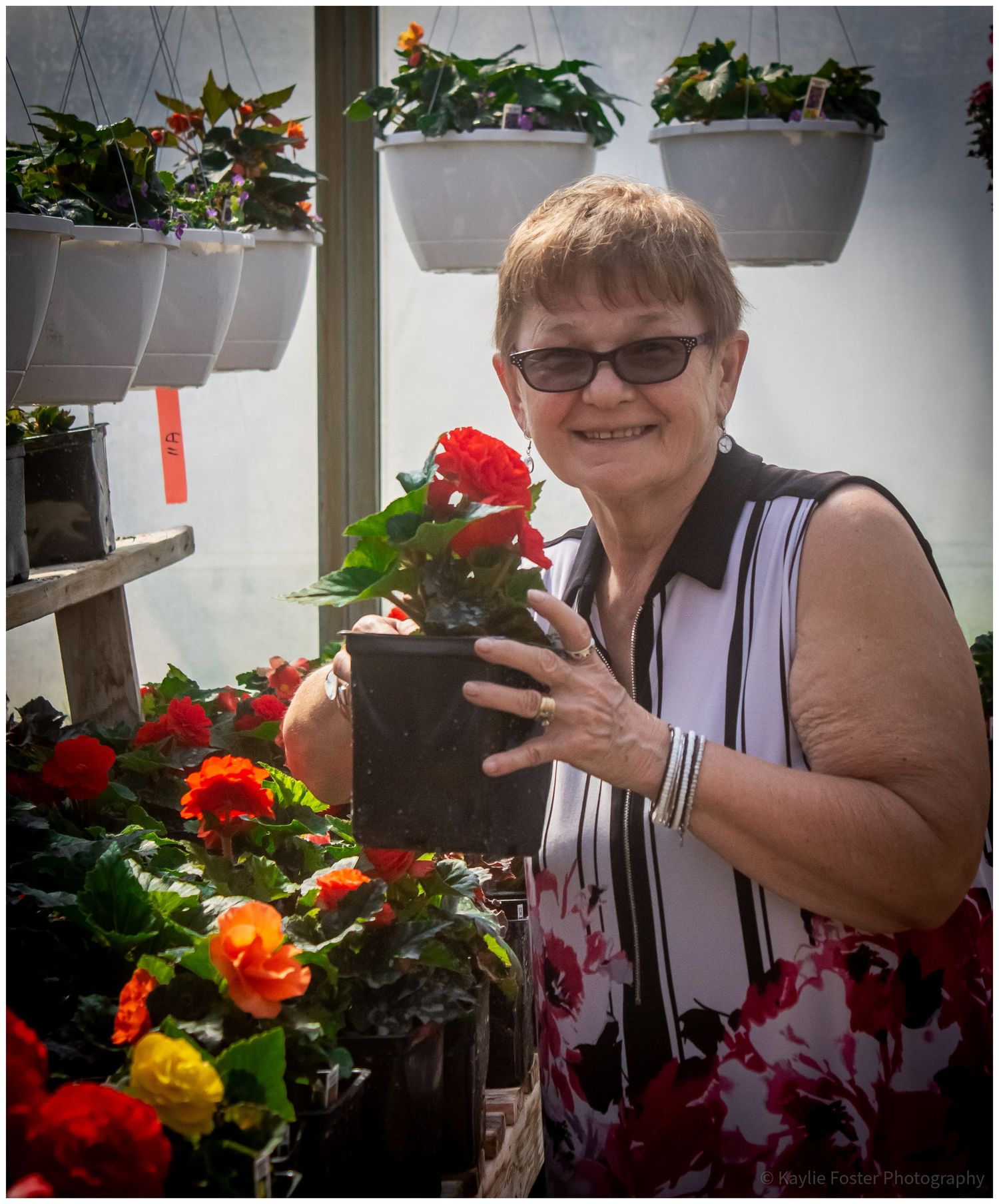 Smiling woman holding a red begonia plant in a greenhouse surrounded by colorful flowers.