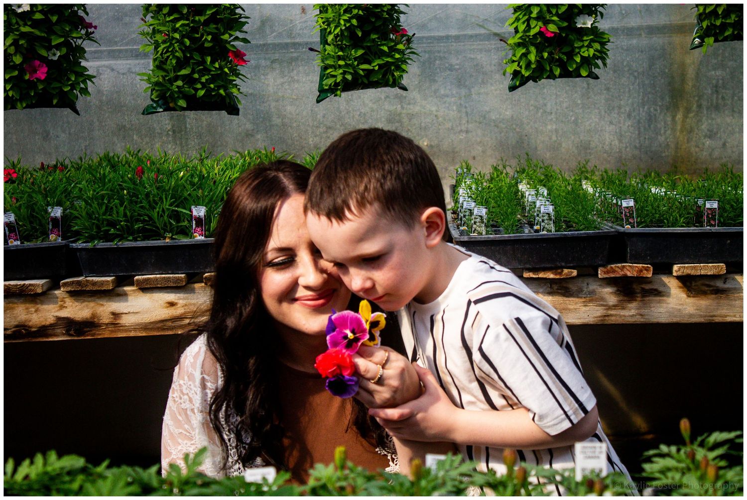 A mother and young boy smell colorful flowers together in a lush garden greenhouse filled with vibrant blooms.
