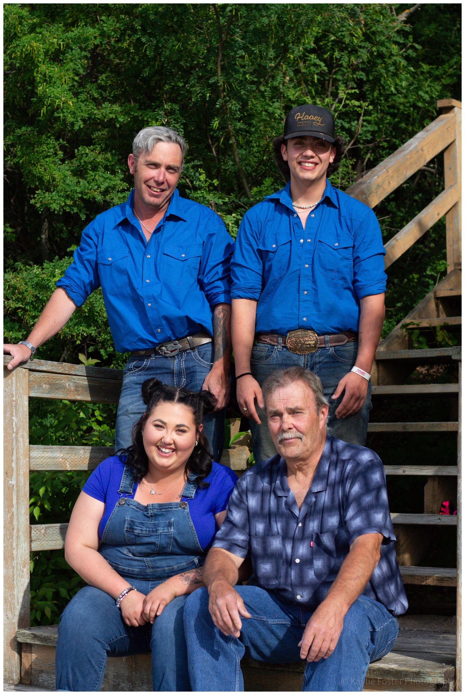 Family of four posing on wooden steps outdoors, two men in blue shirts, woman in overalls, older man in plaid shirt.