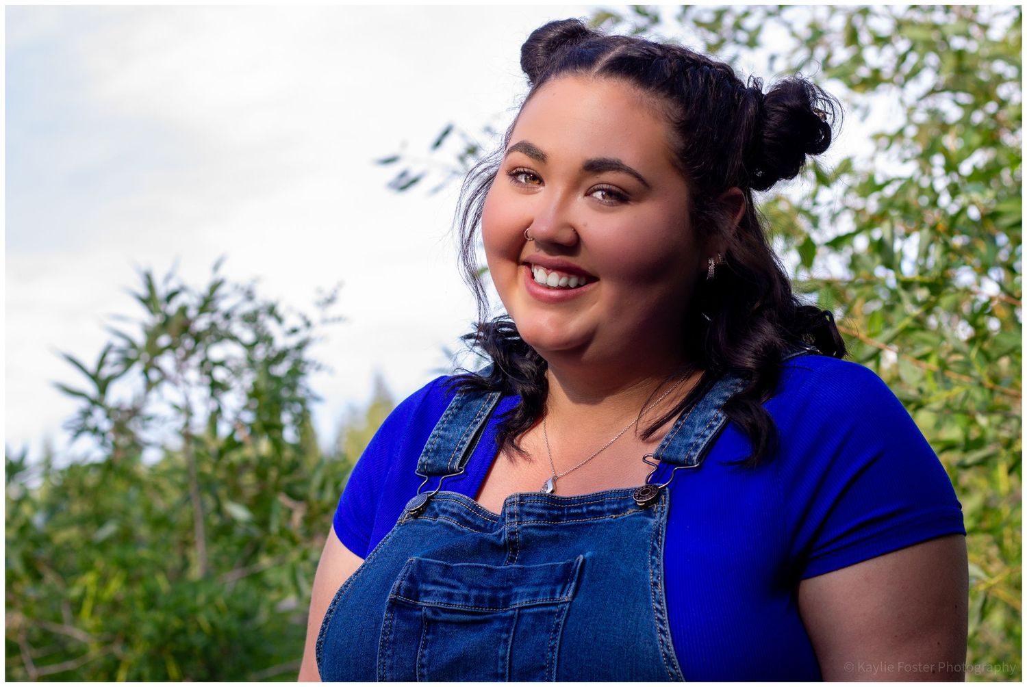 Smiling young woman in blue top and denim overalls outdoors with green foliage background.