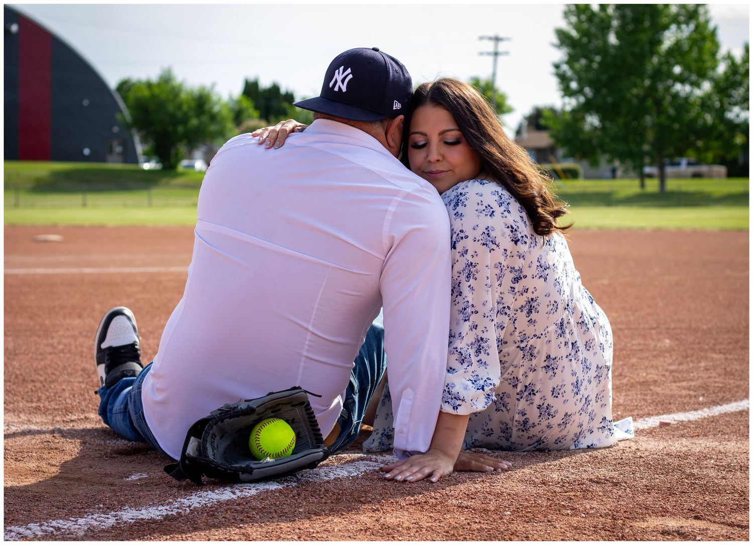 Couple sitting together on a baseball diamond, leaning heads together with a softball and glove nearby.