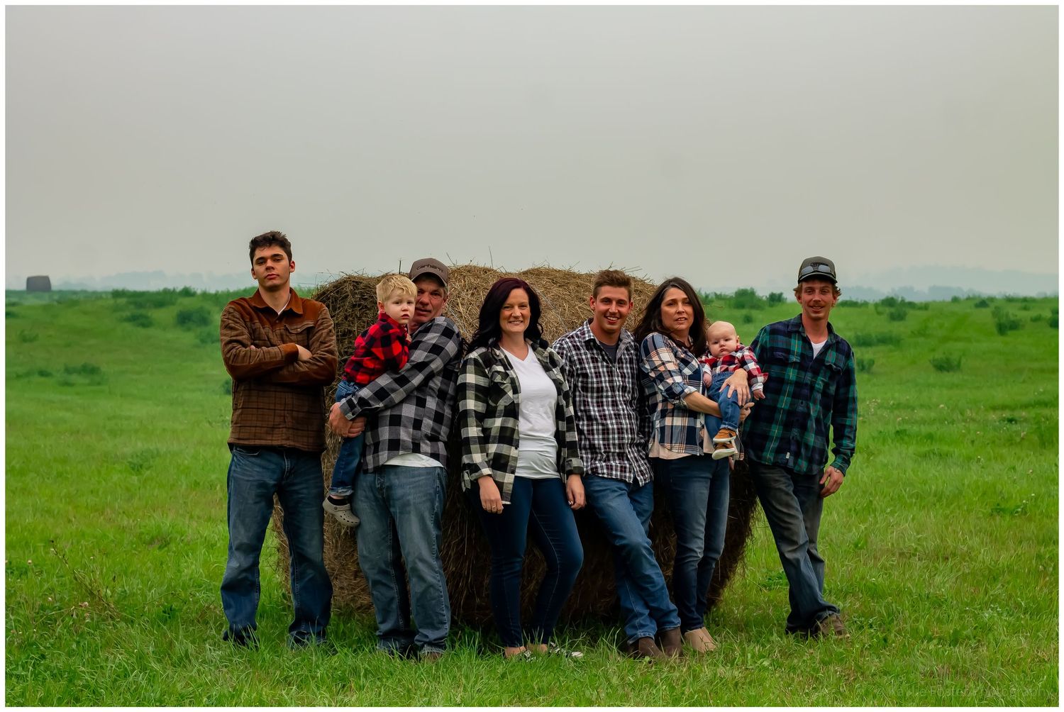 Extended family of seven poses together beside a large hay bale in a green field.