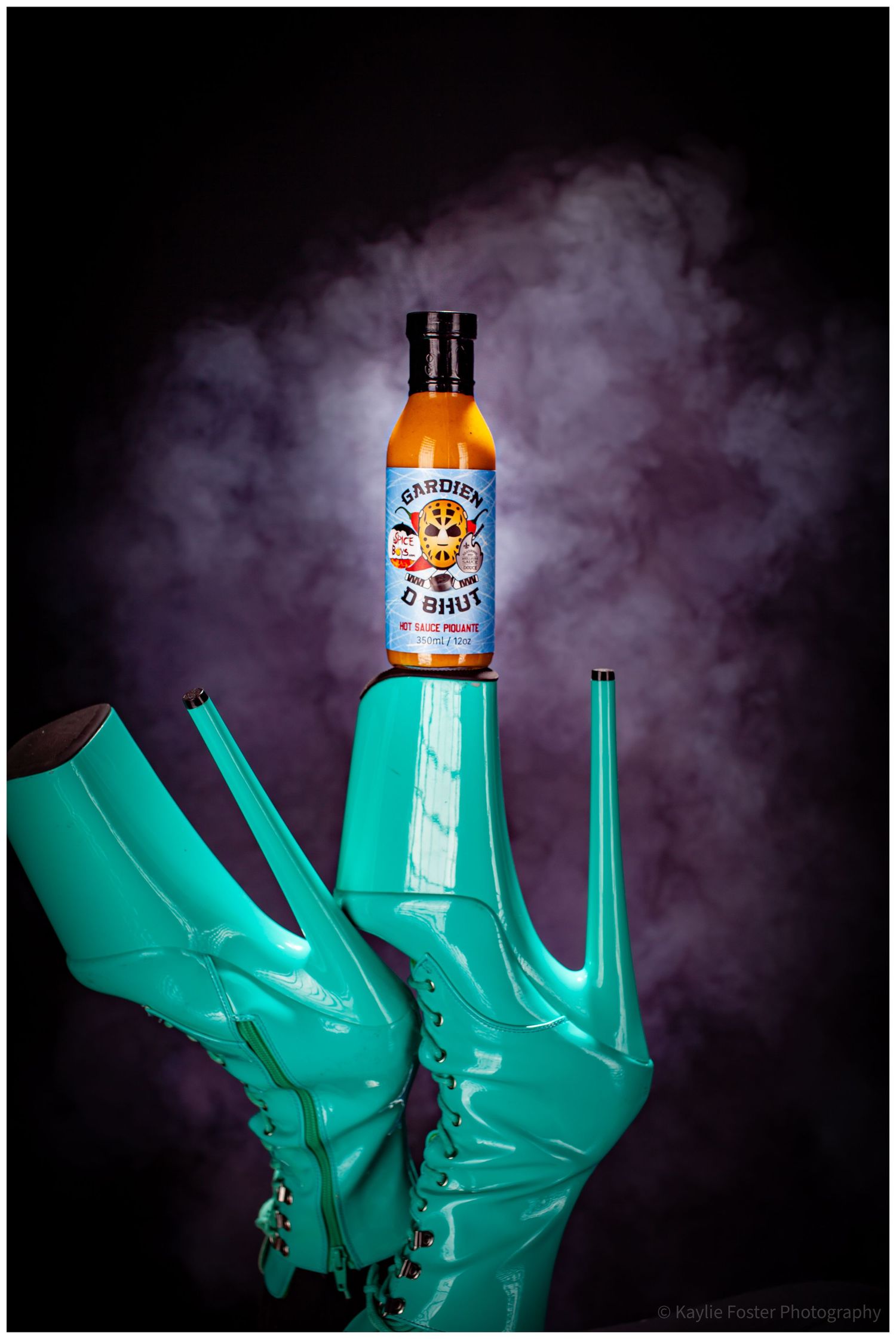 Teal platform boots balancing Gardien D Bhut hot sauce bottle against purple smoky backdrop.