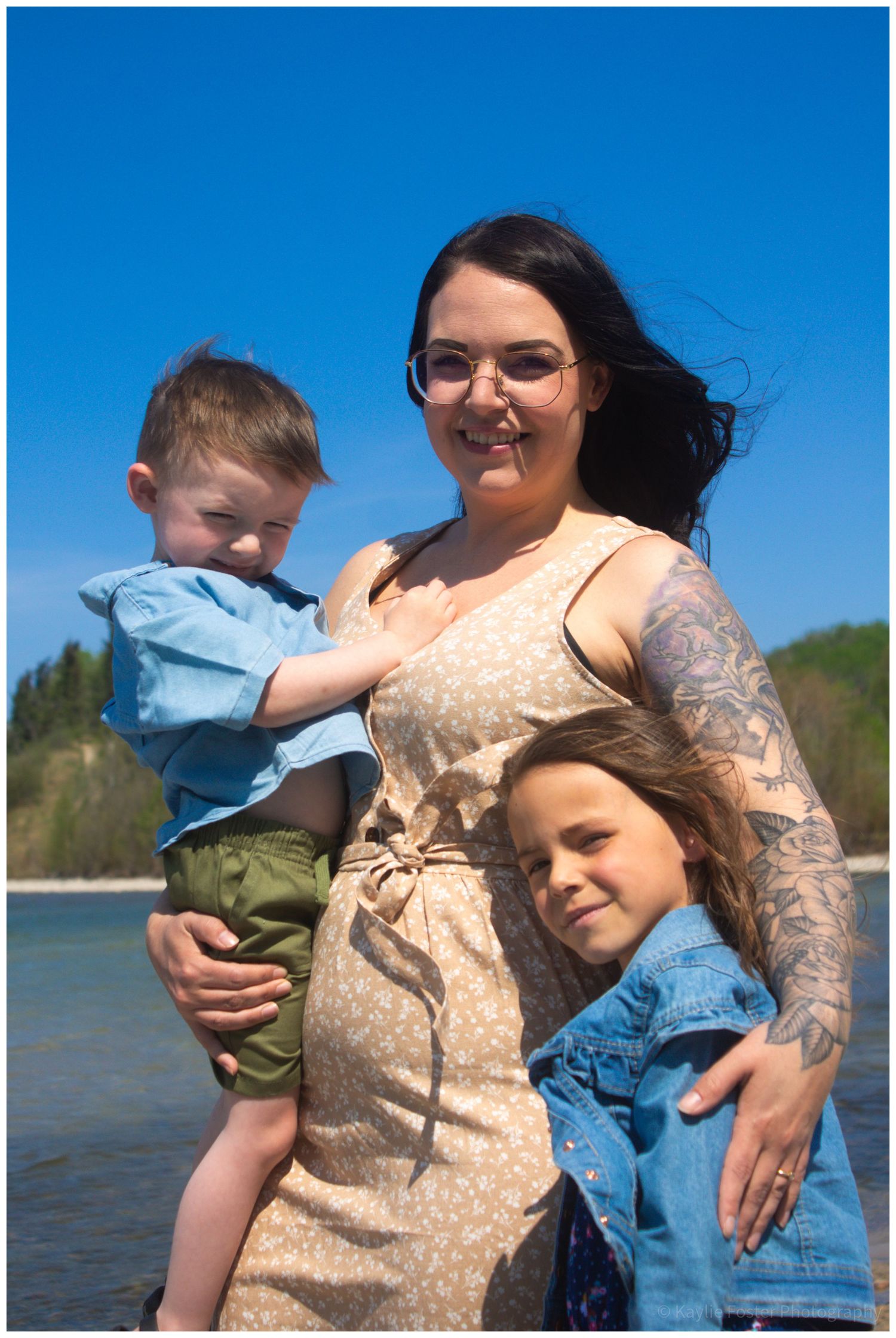 woman with tattoos holds young boy while older child hugs her at a sunny riverside.