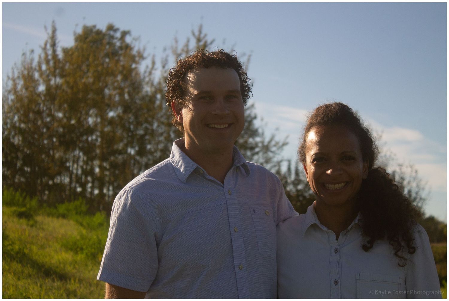 A smiling man and woman standing outdoors on a sunny day with green trees and grass in the background.