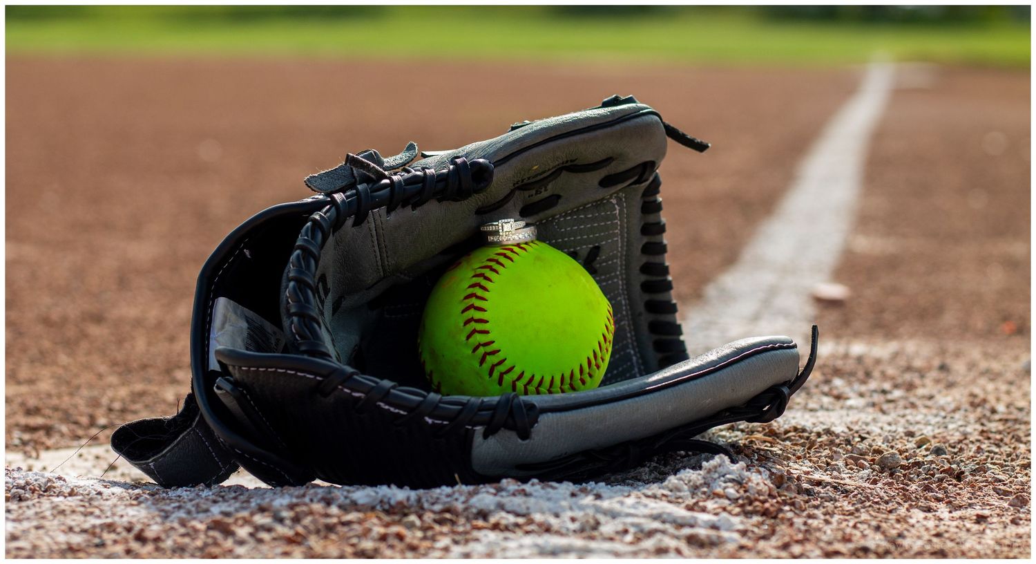 Yellow softball with rings on top resting in a black leather glove on a dirt baseball field near the foul line.