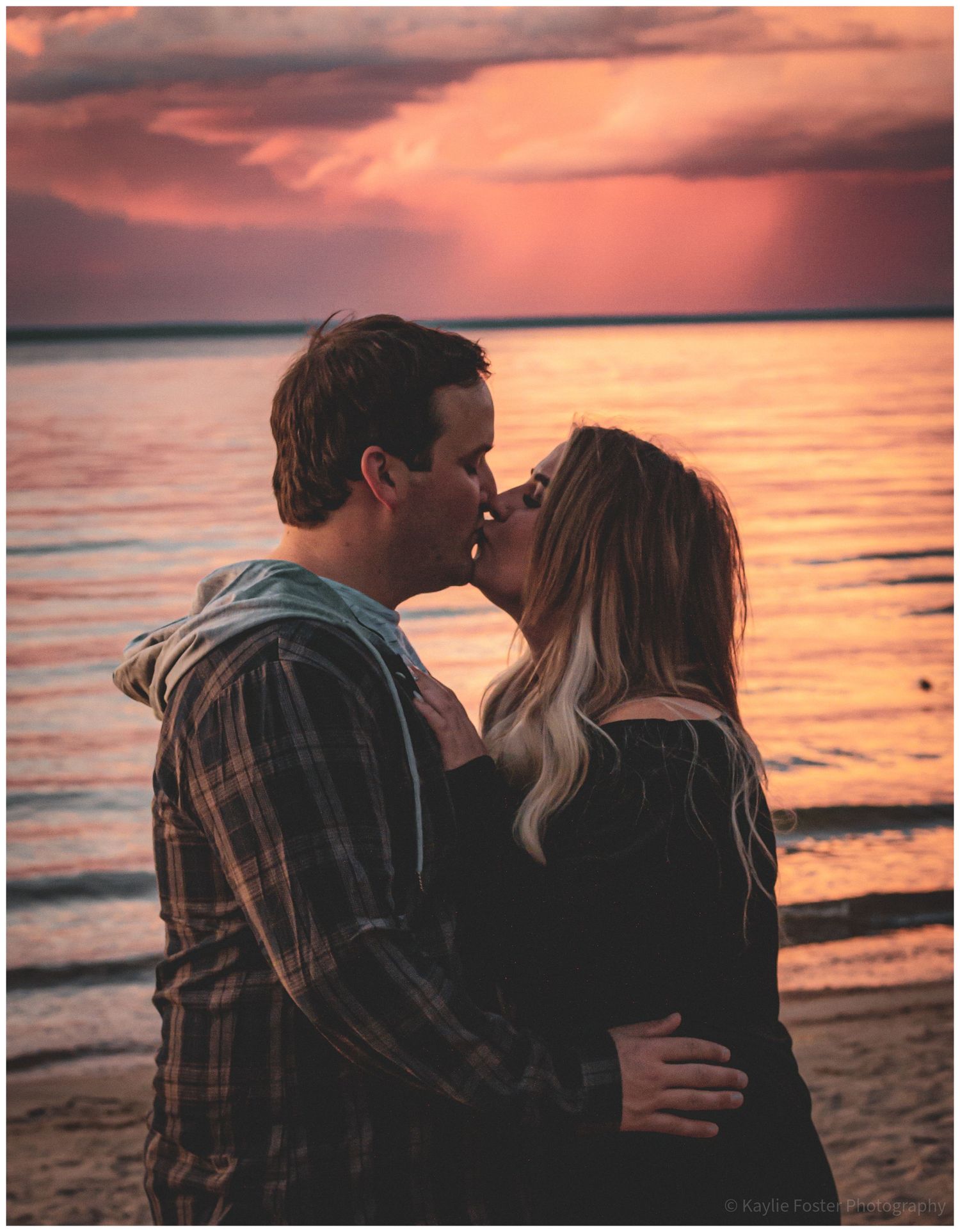 Couple sharing a romantic kiss on the beach at sunset with vibrant pink and orange sky reflecting on calm water.