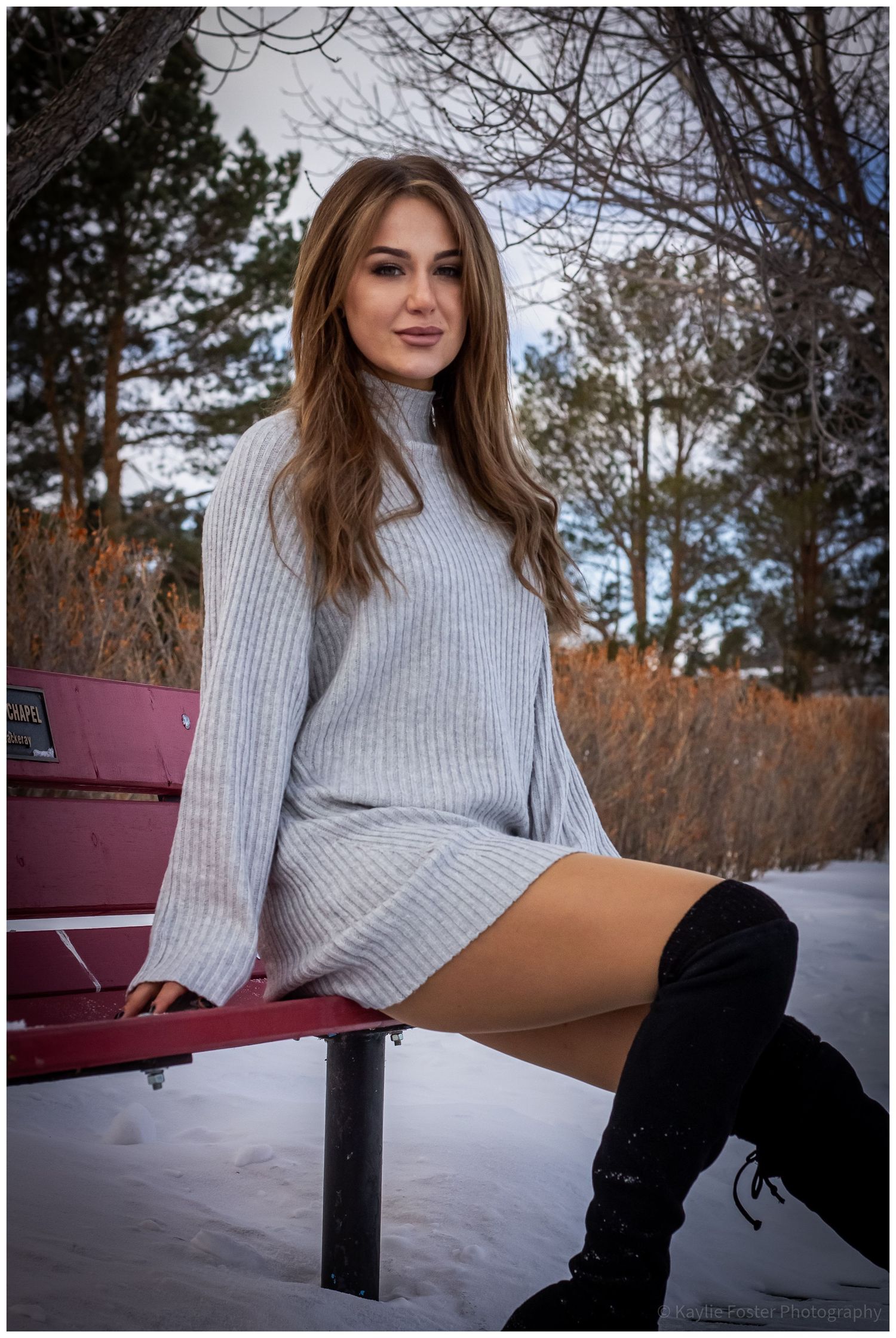 Beautiful woman in gray sweater dress and black thigh-high boots sitting on a red bench in snowy winter park.