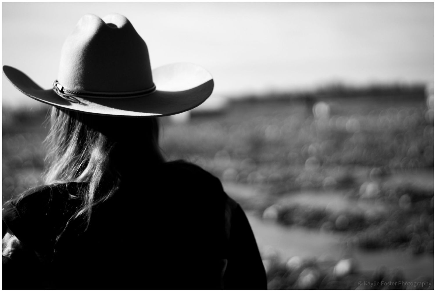 Black and white photo of a woman wearing a cowboy hat, viewed from behind, overlooking a rural landscape.