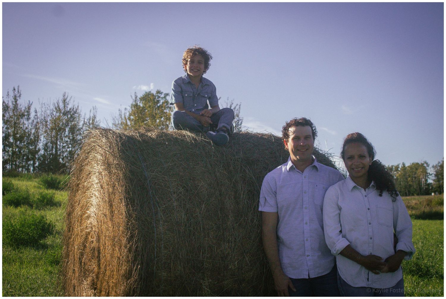 Family of three posing by a large round hay bale in a sunny green field, child sitting on top.