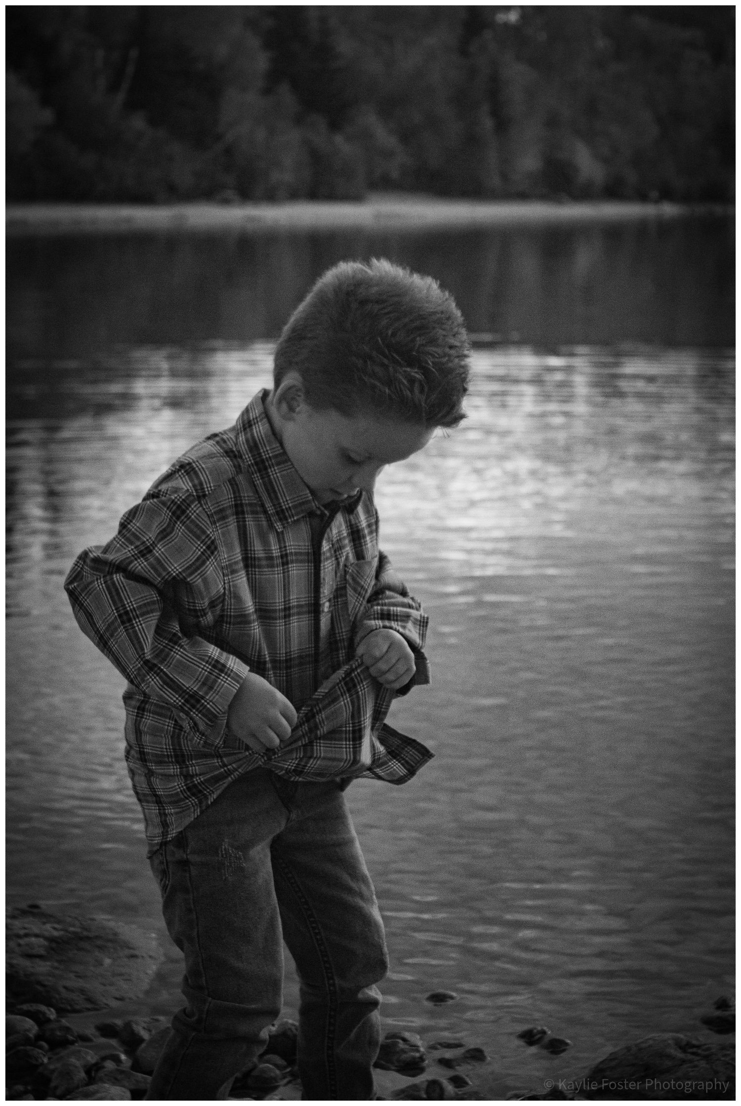 Black and white photo of a young boy standing by a lake adjusting his plaid shirt.