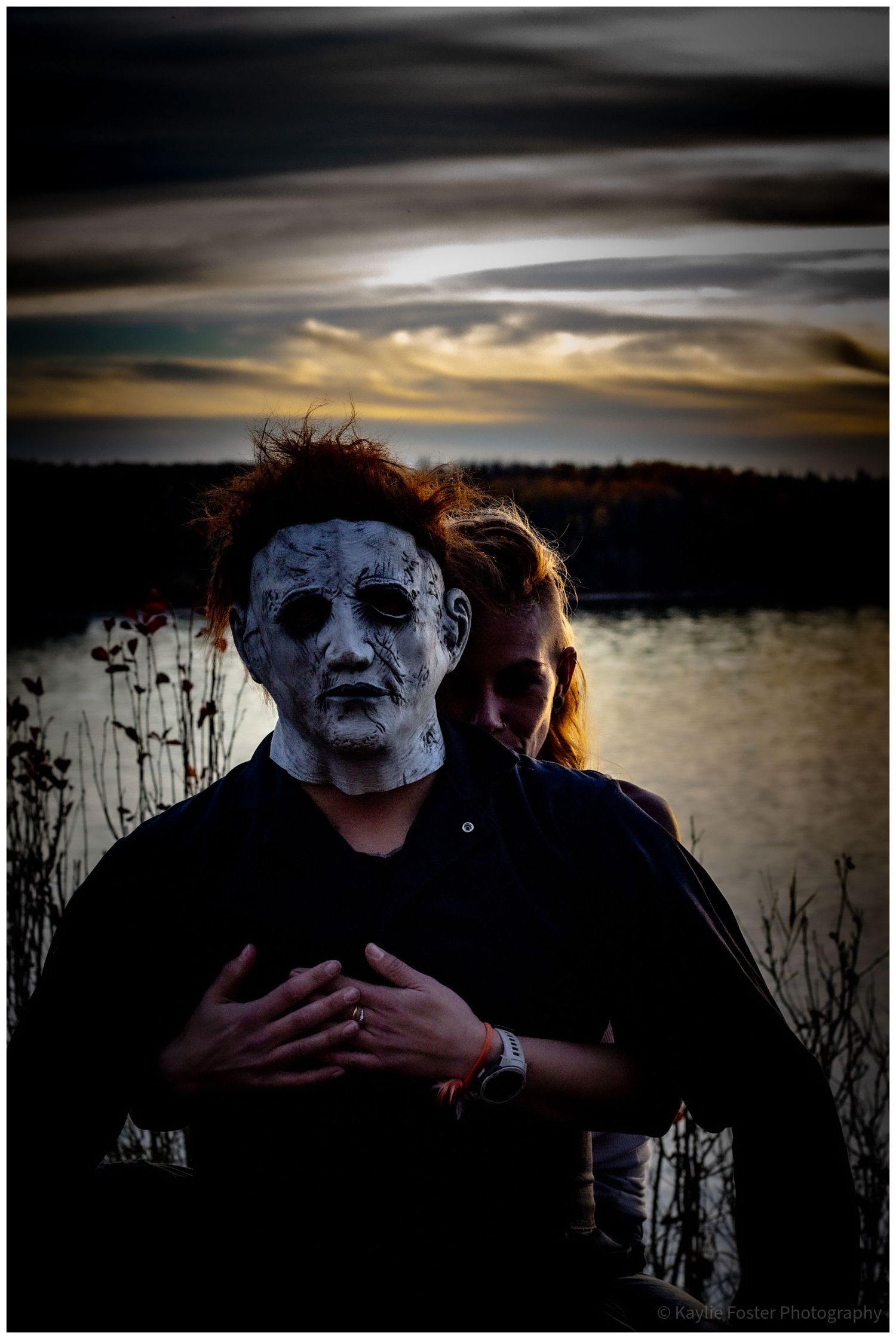 Person wearing Michael Myers Halloween mask embraced by woman near lake at dramatic sunset.