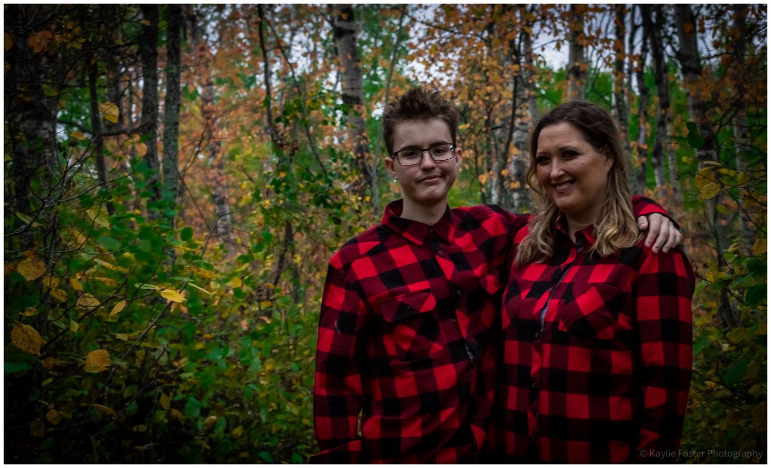 Mother and teenage son in matching red plaid shirts in autumn forest.