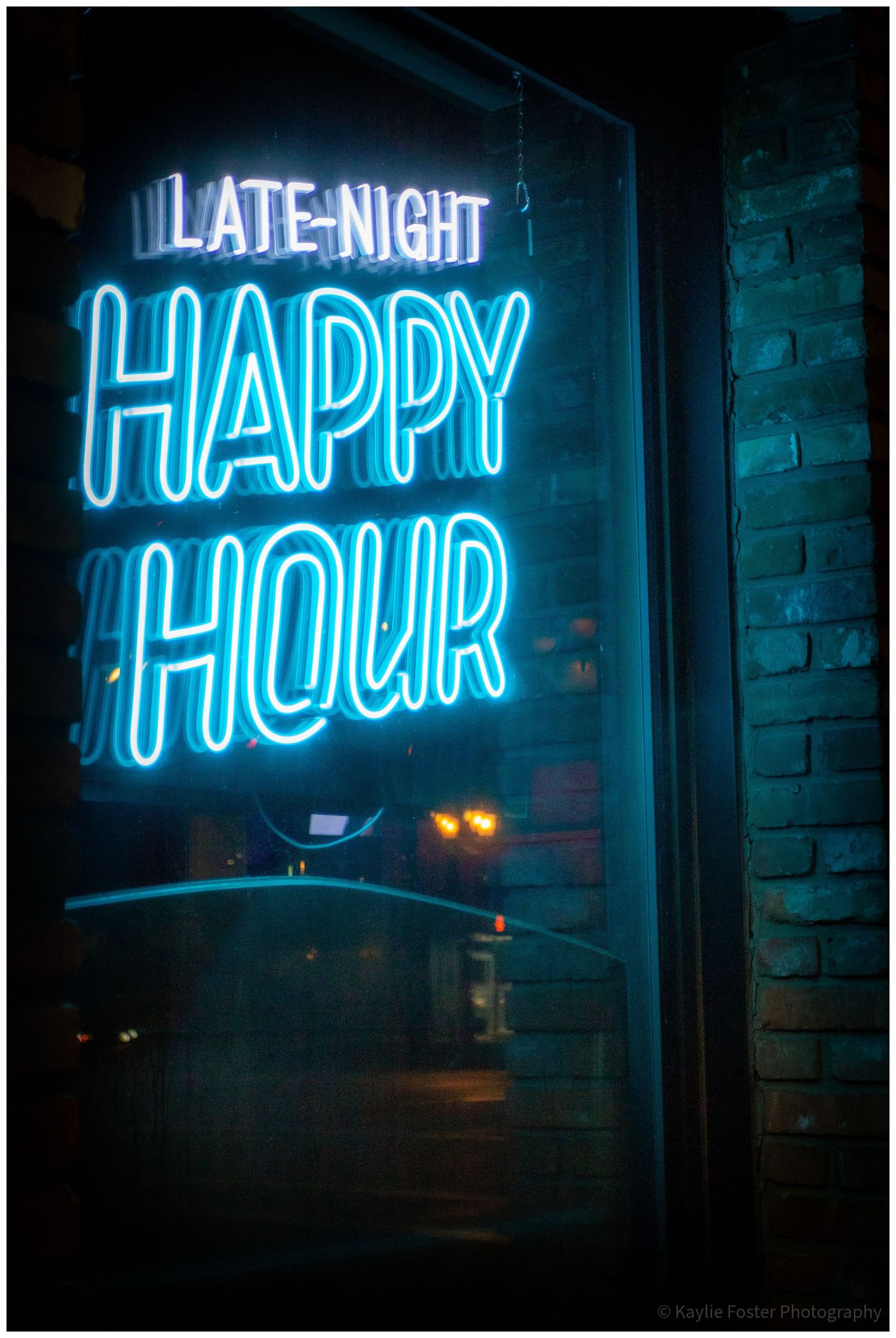 Glowing blue neon sign reading Late Night Happy Hour in a dark bar window with brick wall backdrop.