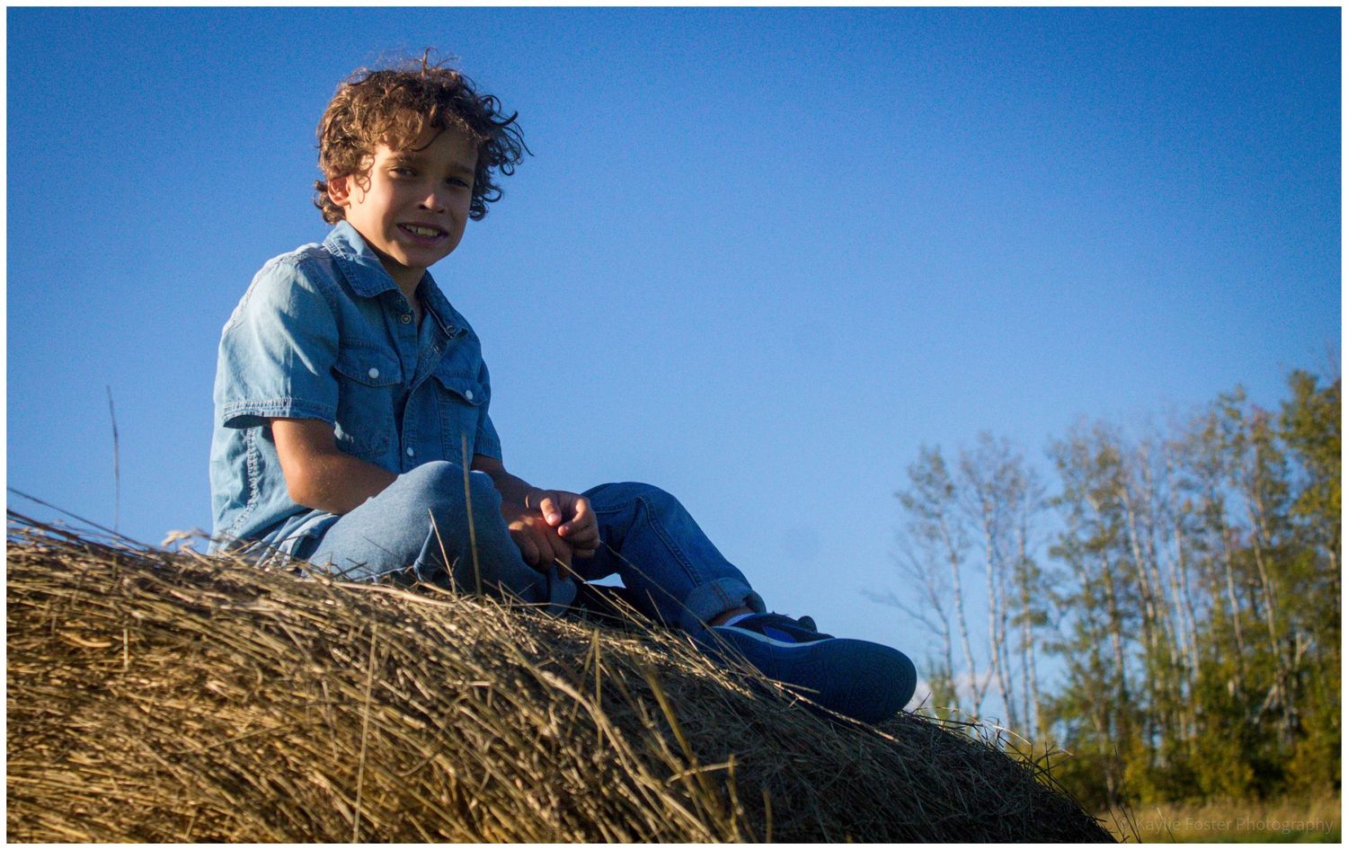 A curly-haired boy in denim sits atop a hay bale under a bright blue sky.