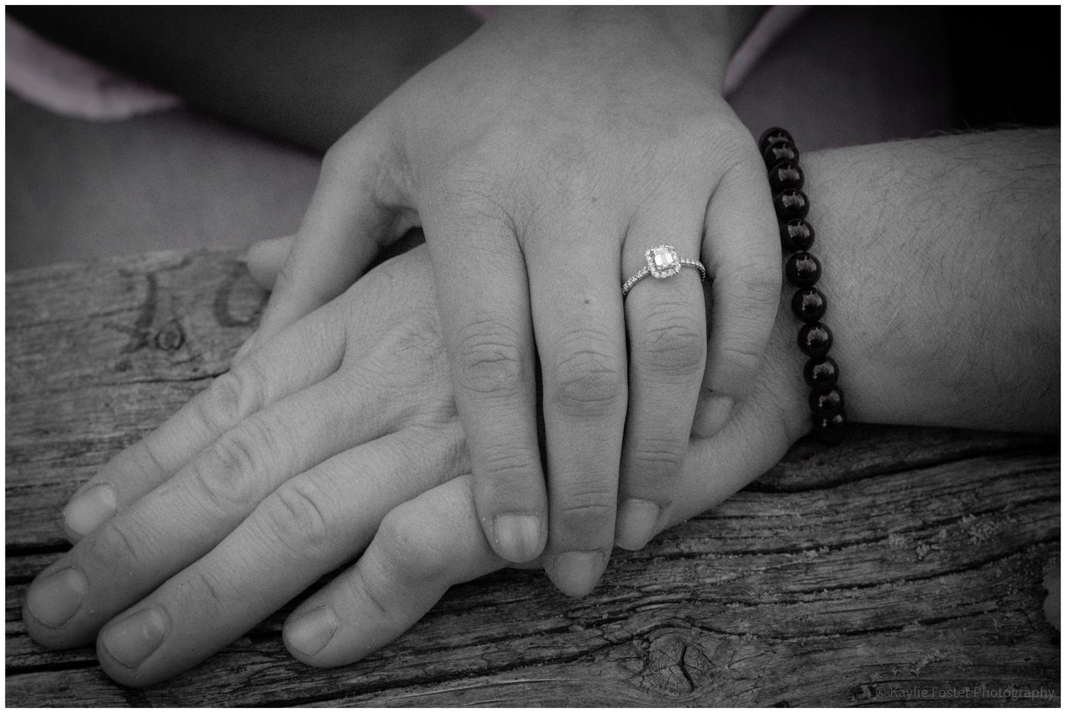 Black and white photo of two hands touching, one wearing an engagement ring and the other a beaded bracelet, on wood.
