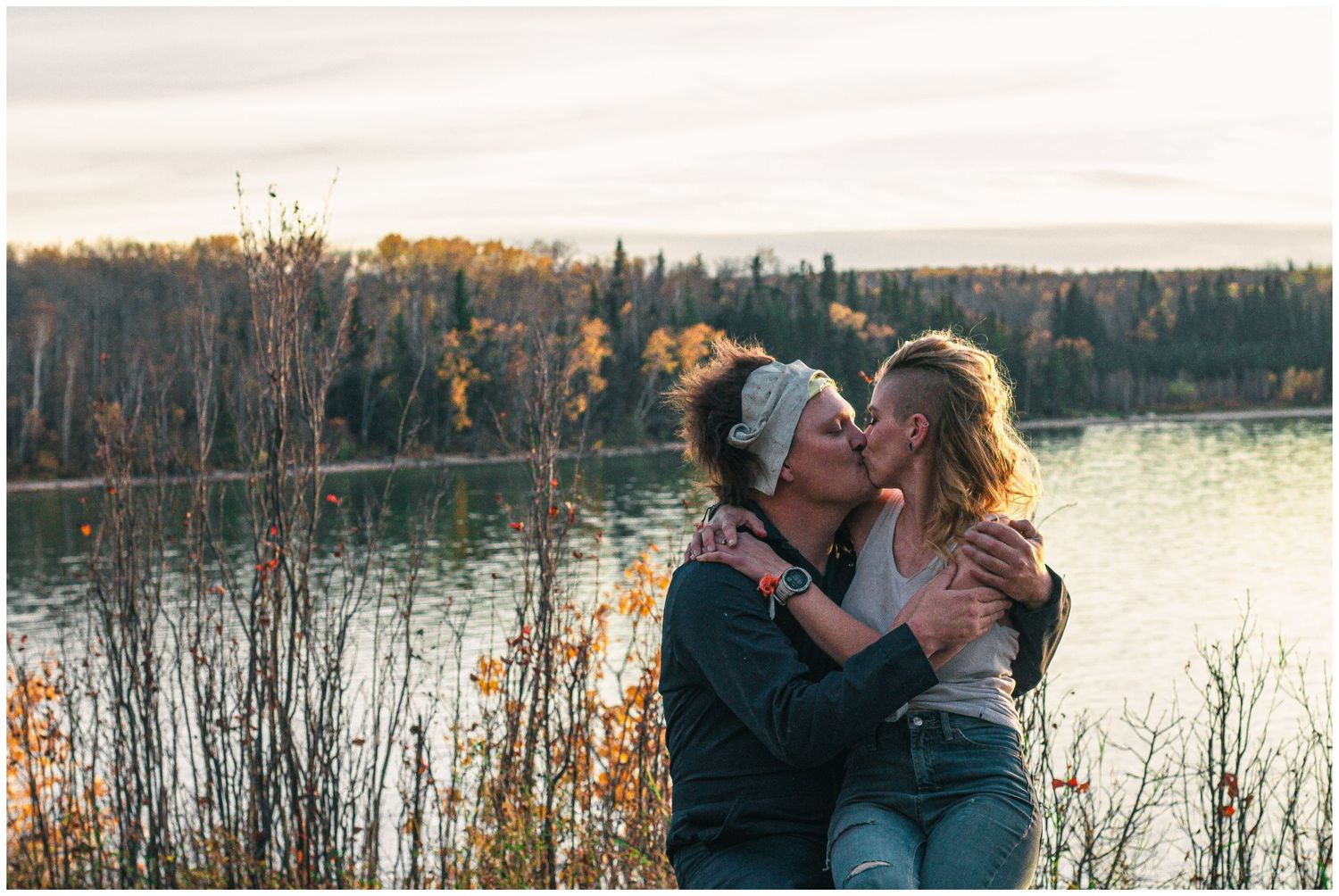 Couple sharing a romantic kiss outdoors by a serene lake surrounded by colorful autumn foliage at golden hour.