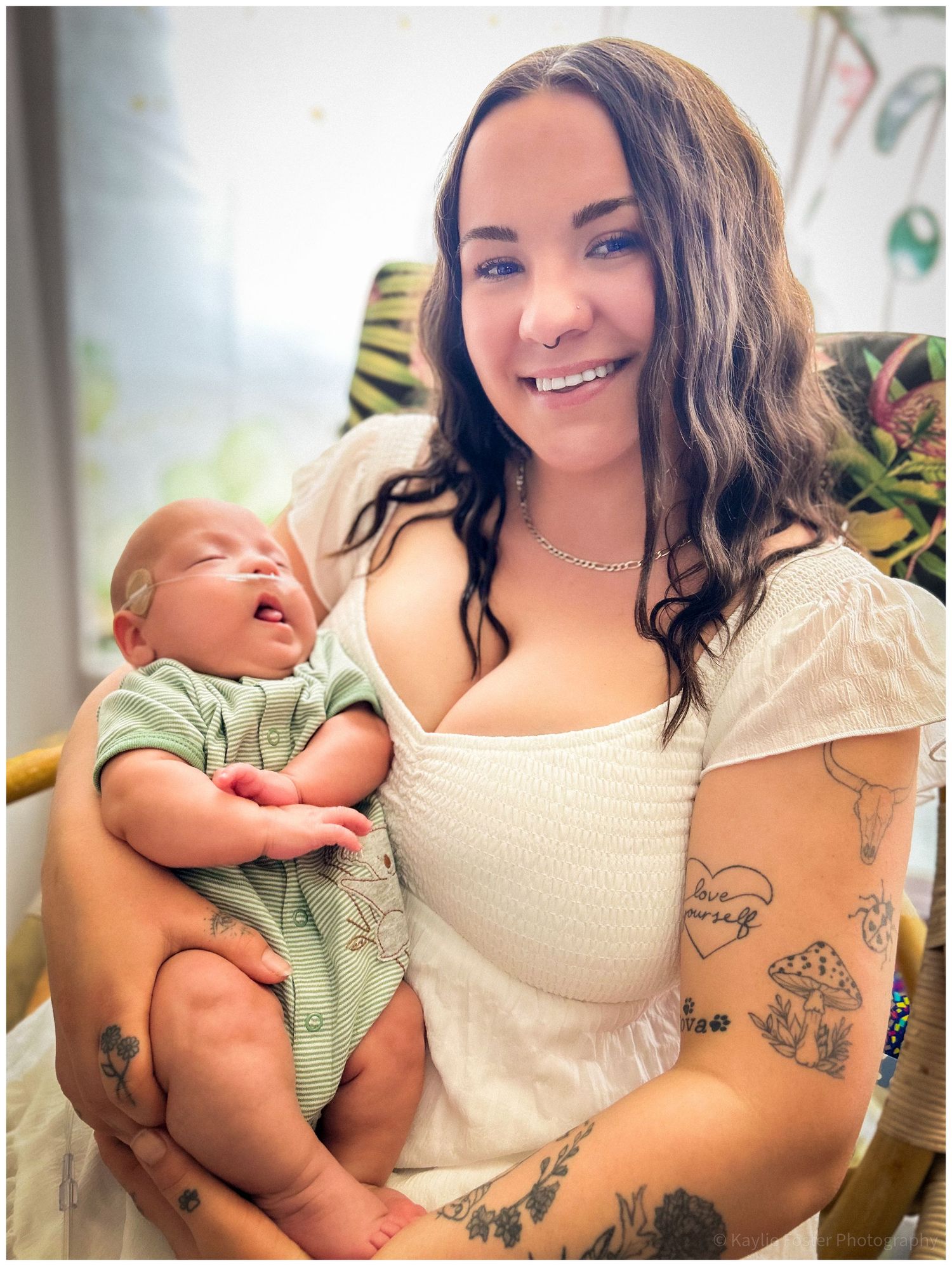 Smiling young woman with tattoos holding a sleeping newborn baby in a green onesie indoors.