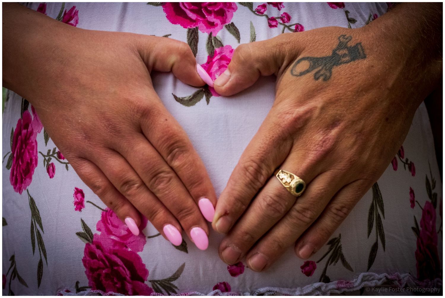 Two hands forming a heart shape on a pregnant belly, one wearing a gold ring and pink nail polish, over a floral dress.