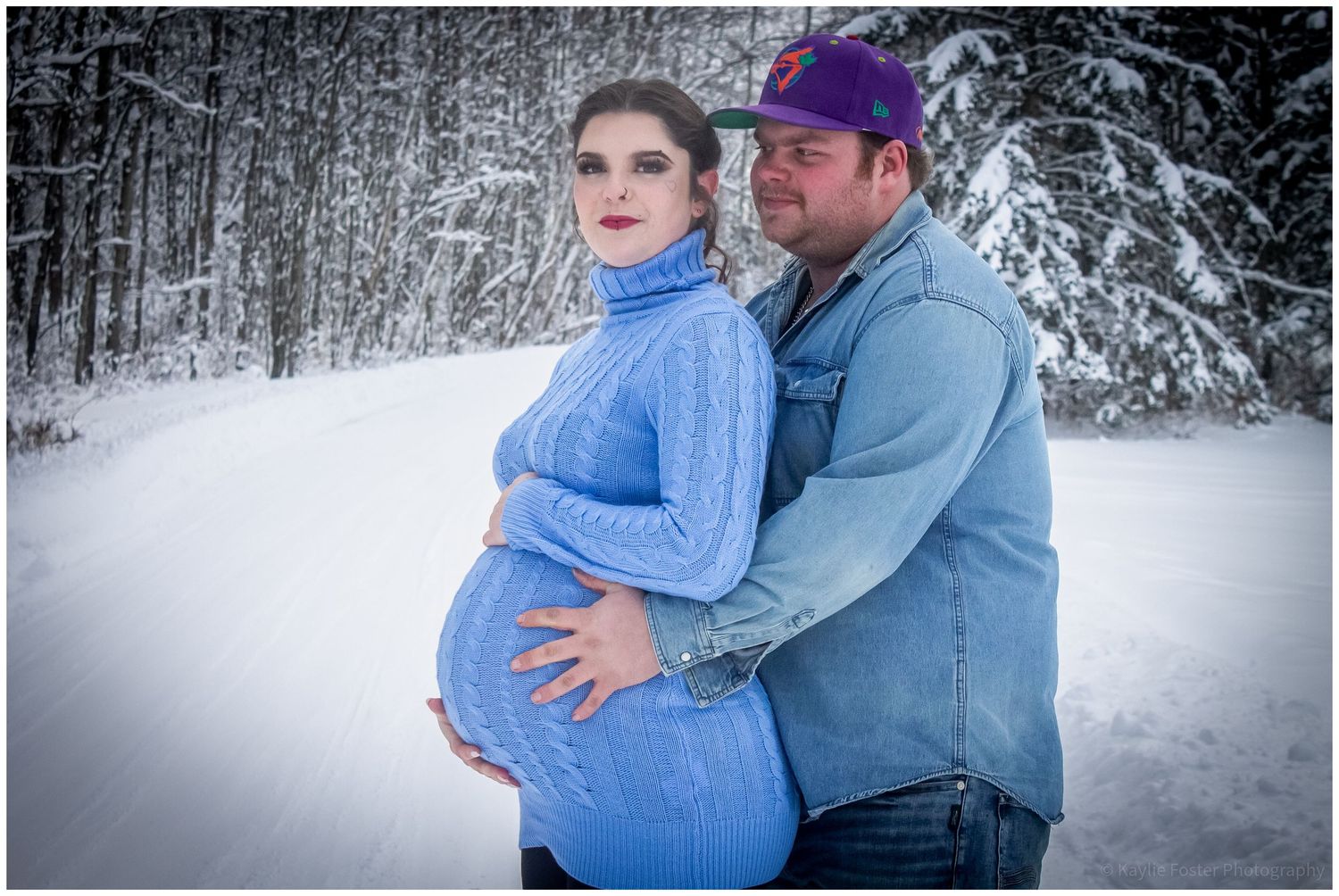 Pregnant couple poses in snowy winter forest, woman in blue sweater dress, man in denim jacket and purple cap.