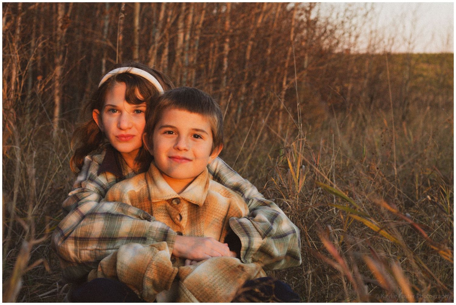 Two children in plaid jackets sit together in a golden field at sunset, the older girl hugging the younger boy.