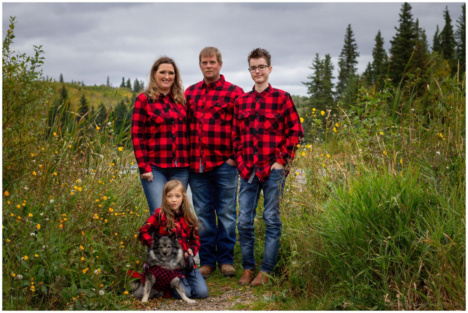 Family of four in matching red plaid shirts pose outdoors with their dog in a meadow.
