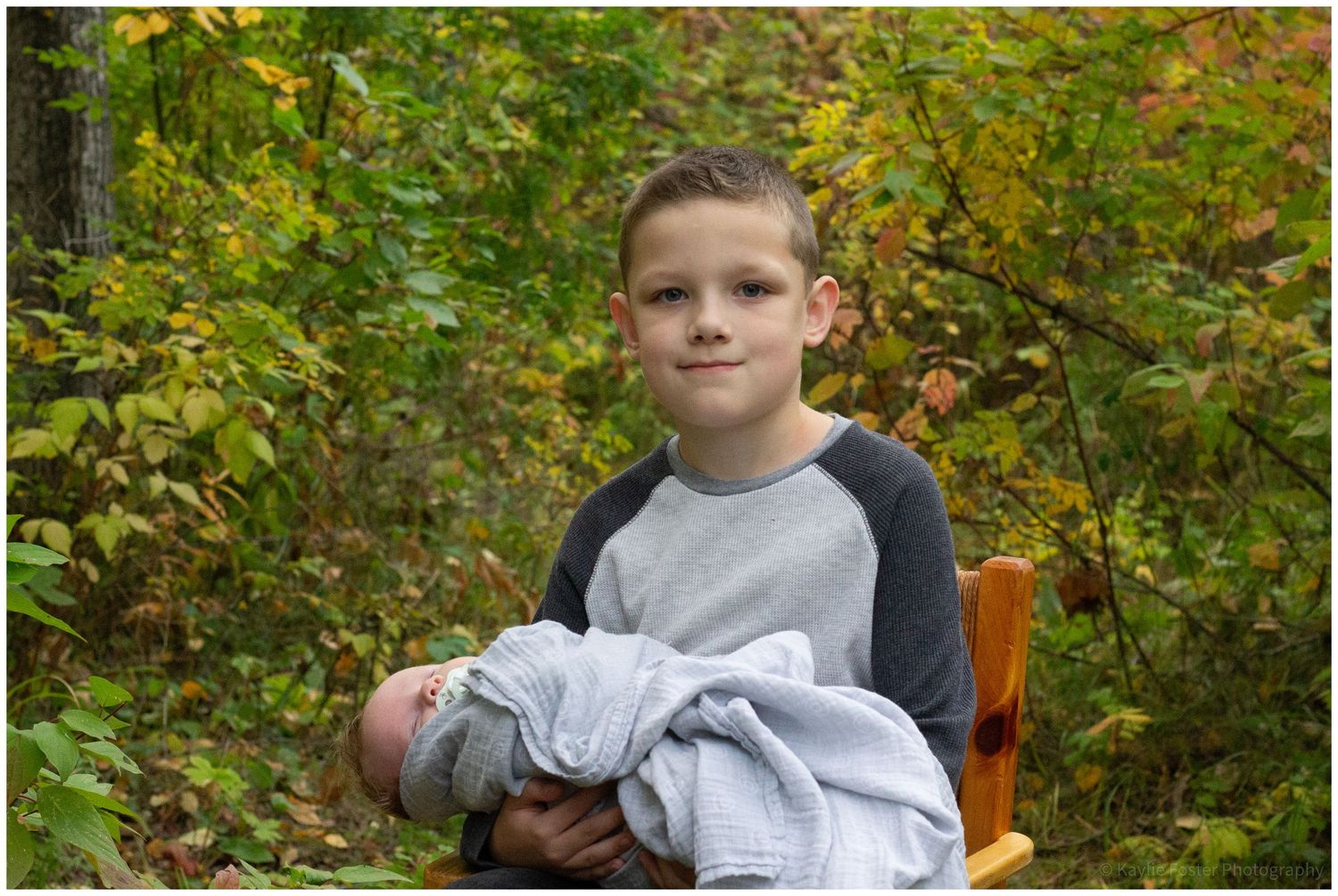 A young boy holds a newborn baby wrapped in a light blue blanket outdoors among autumn foliage.