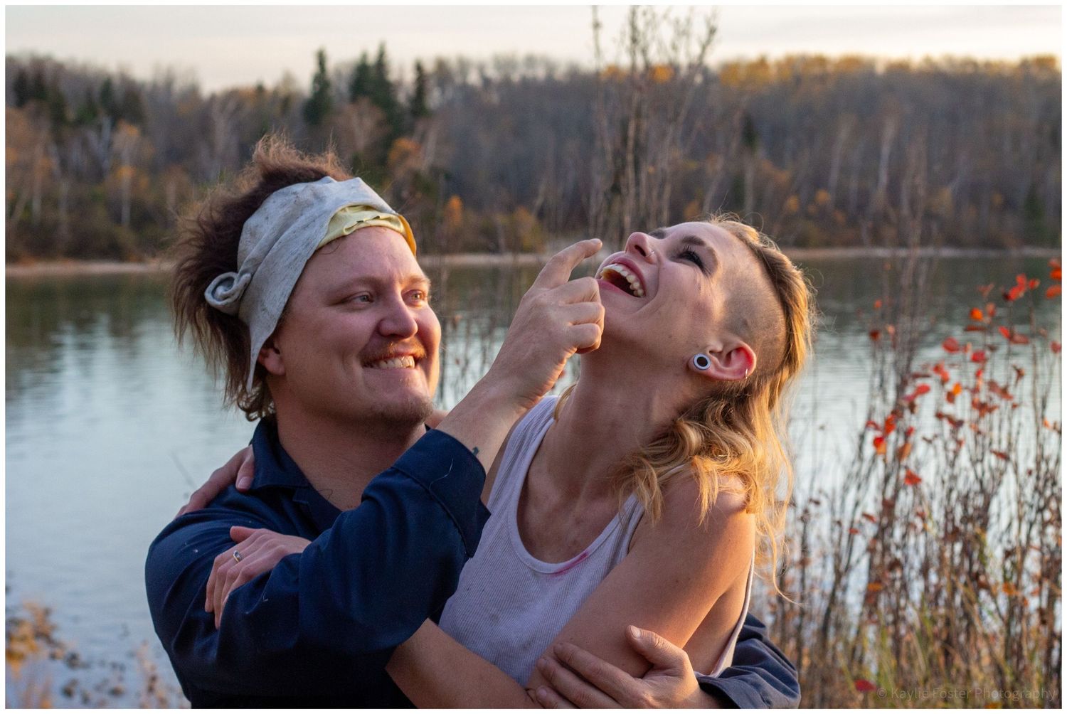 Two women laughing and playing outdoors near a lake surrounded by autumn foliage at sunset.