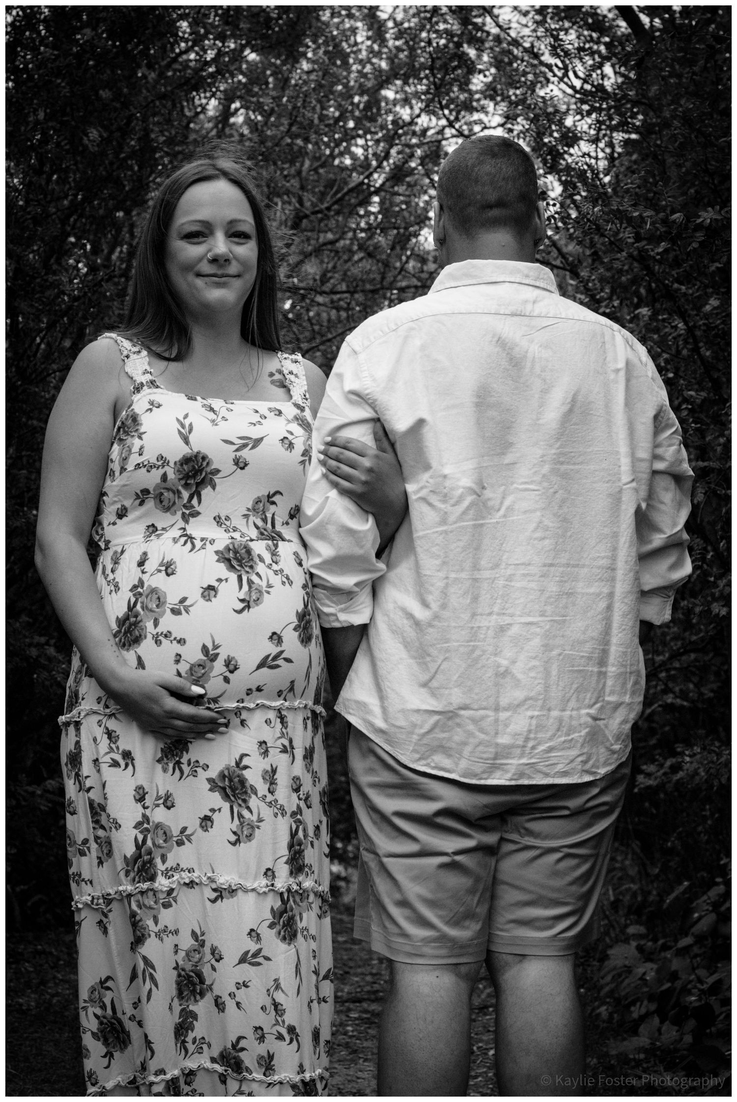 Black and white maternity photo of pregnant woman in floral dress with partner outdoors.
