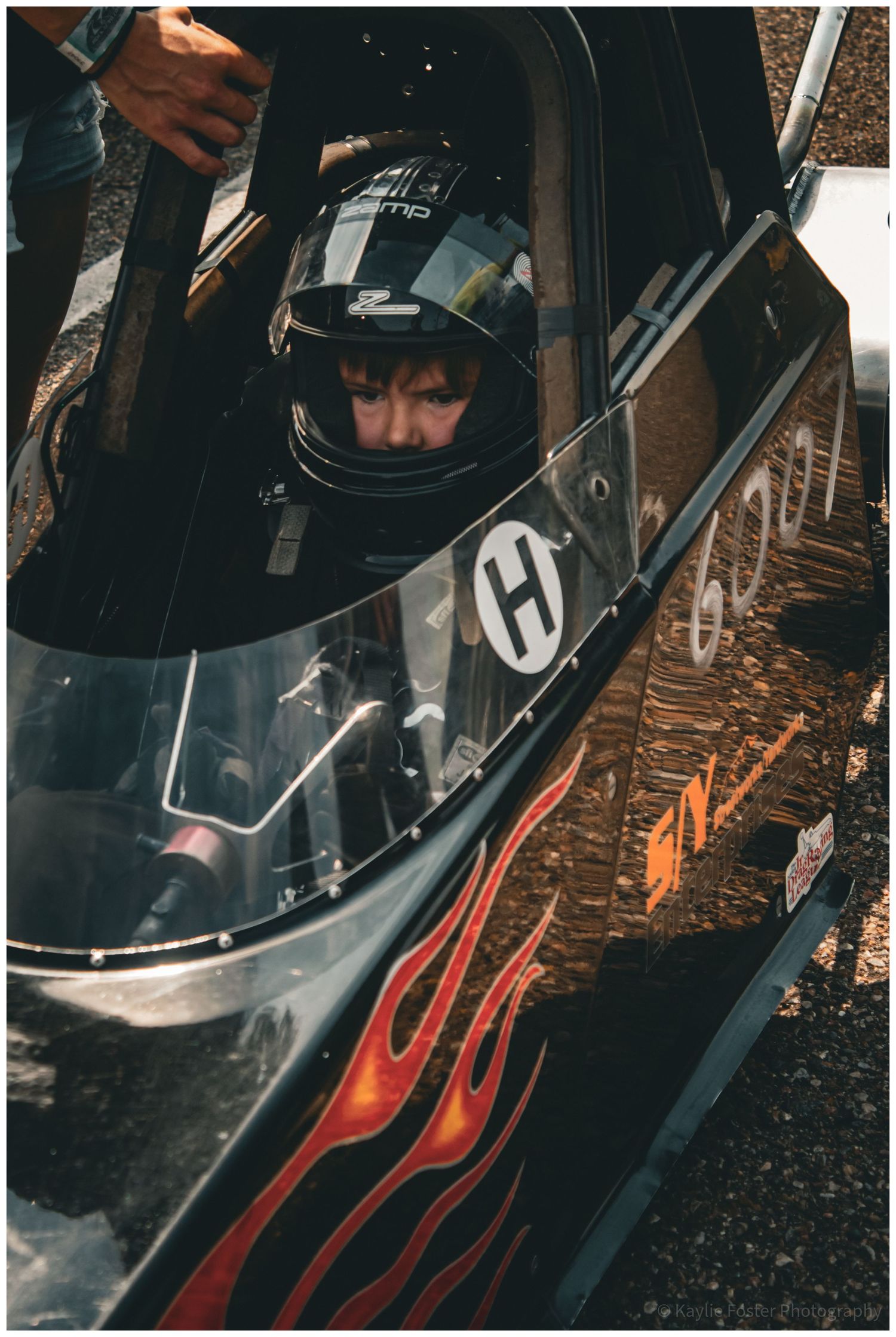 Young driver wearing a helmet seated inside a flame-decorated drag racing car at a race event.