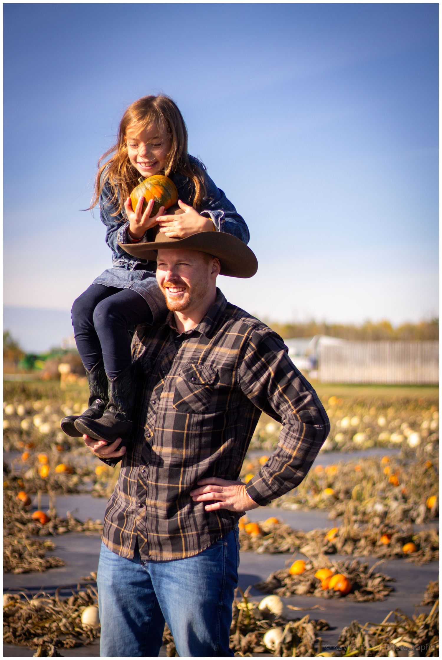 A man in a cowboy hat carries a smiling girl on his shoulders holding a pumpkin in a sunny pumpkin patch.