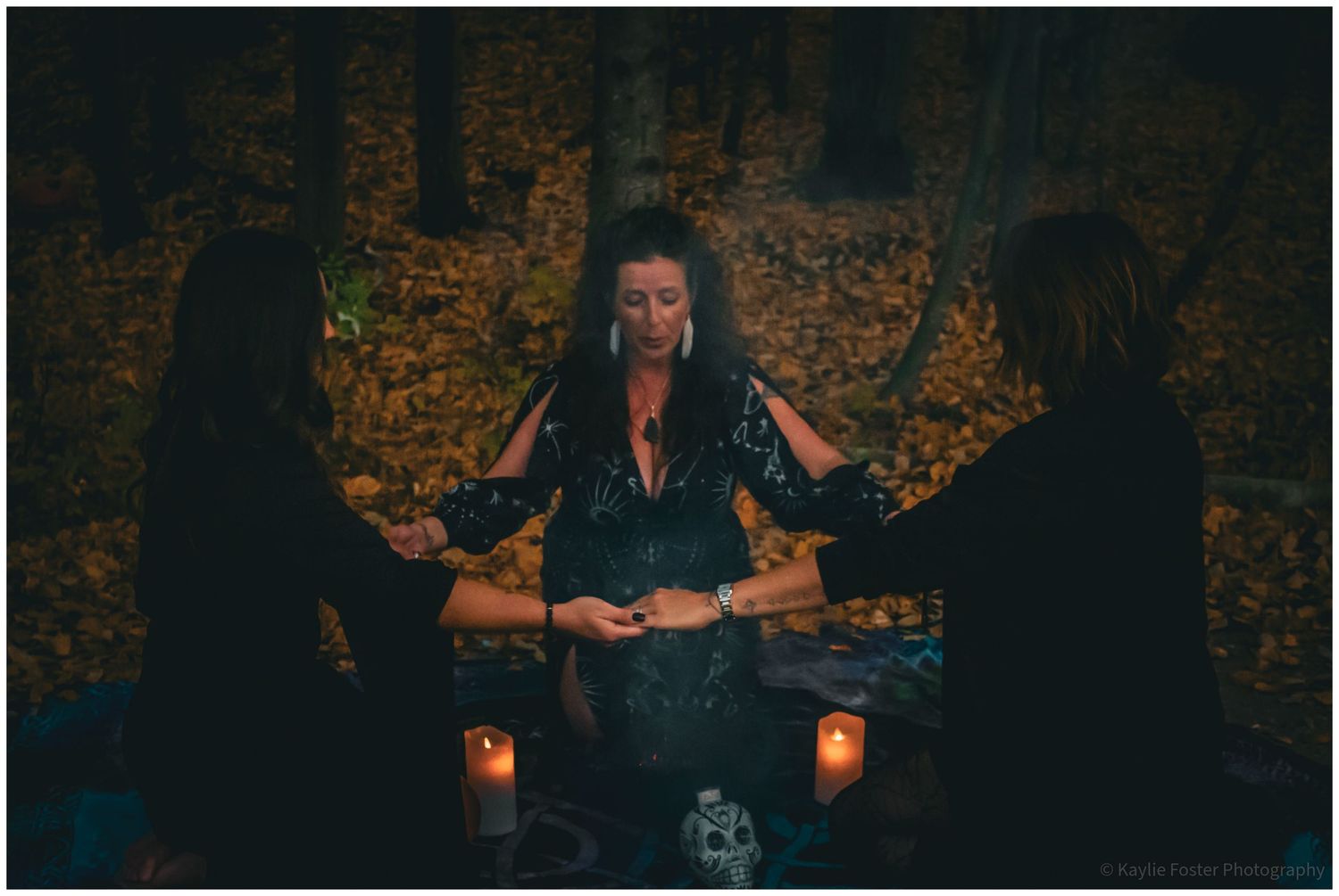 Three people holding hands around candles in a nighttime woodland ritual ceremony setting.