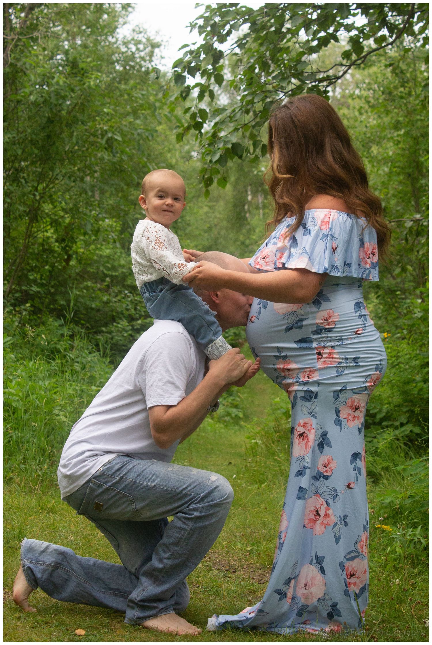 Expectant family maternity photo outdoors, father kissing pregnant belly while holding toddler.