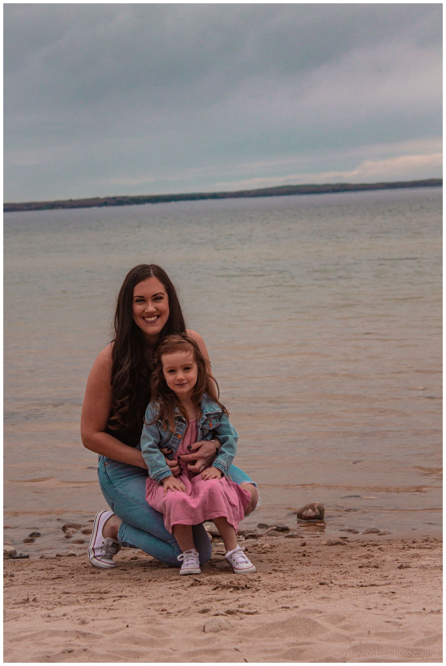Mother and young daughter posing together on a sandy beach by calm water.