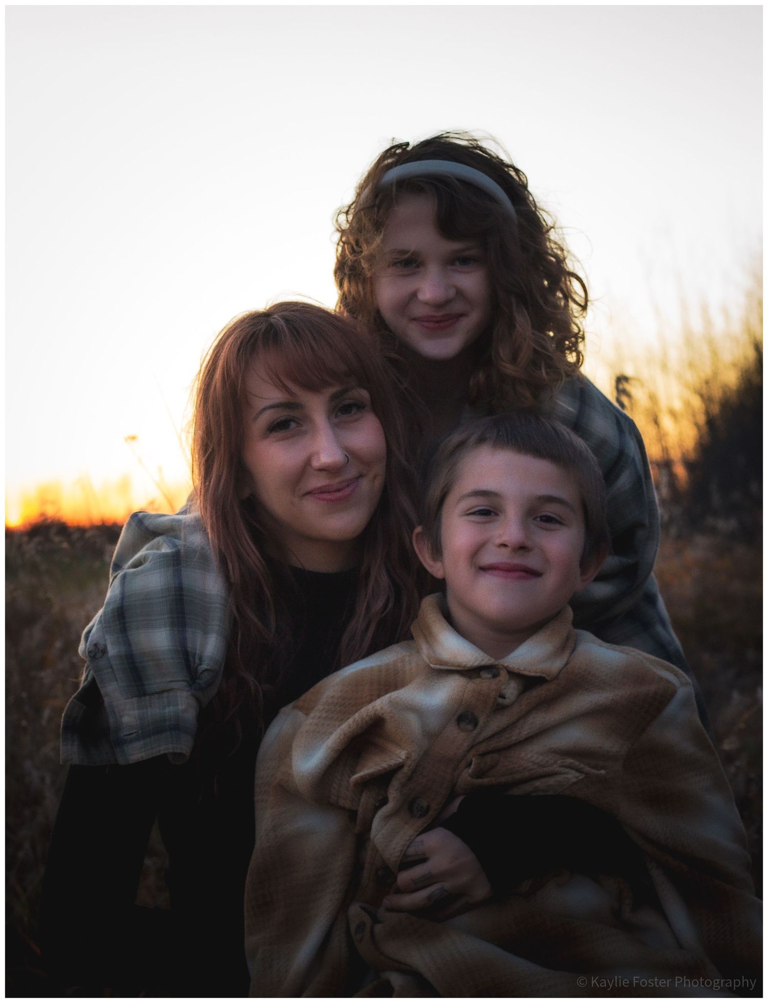 A mother smiles with her two children outdoors at sunset in a field.