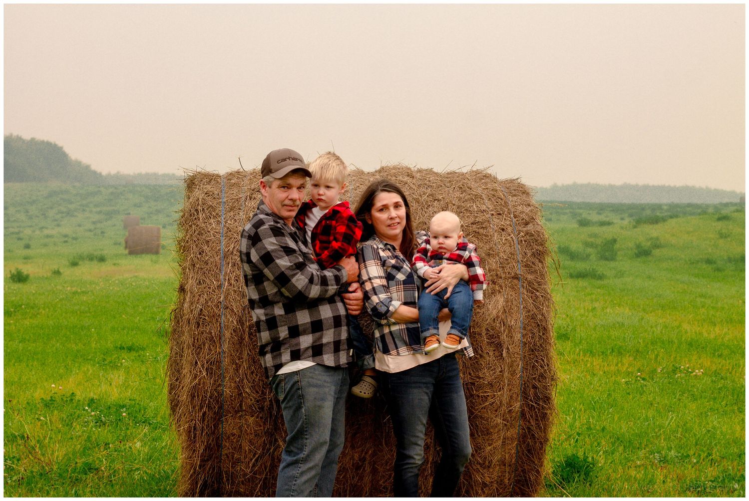 Couple in plaid shirts holds two young children beside a large hay bale in a green field.