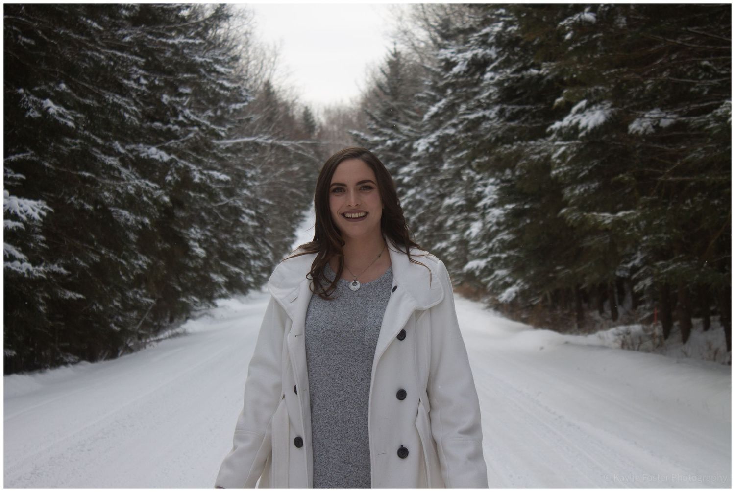 Woman in white coat smiling on snowy tree-lined road in winter.