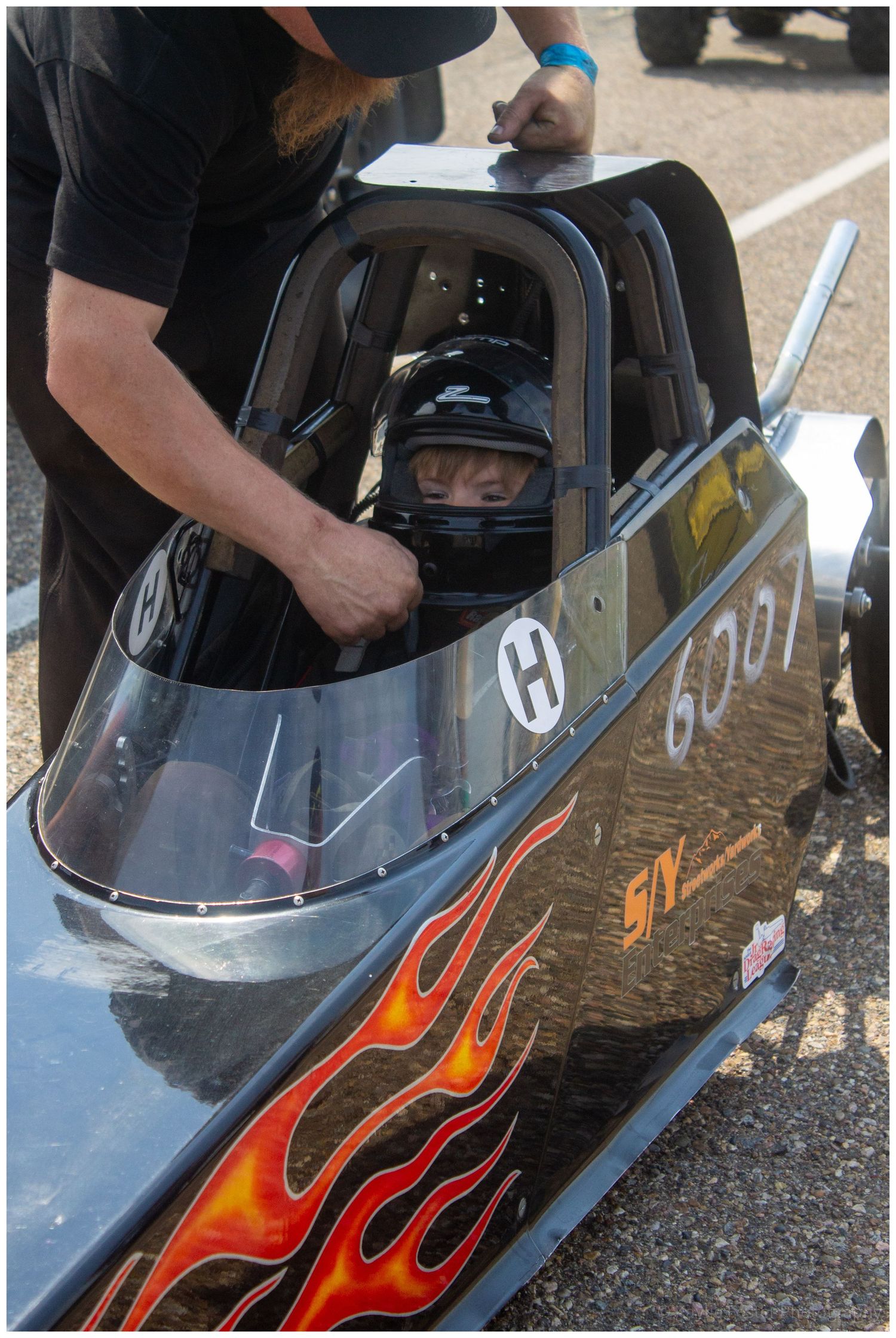 Young driver in helmet seated in flame-decorated dragster number 6001 being assisted by crew.
