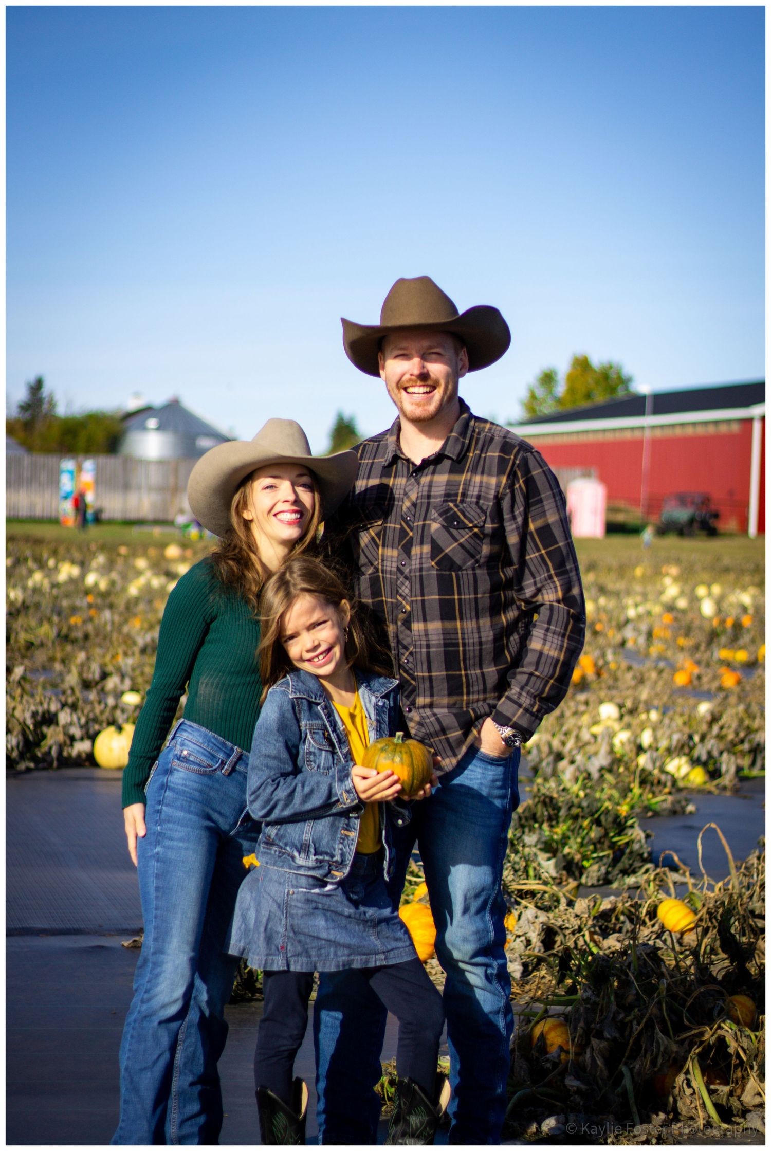 Family of three smiling in a pumpkin patch, wearing cowboy hats and jeans on a sunny fall day.