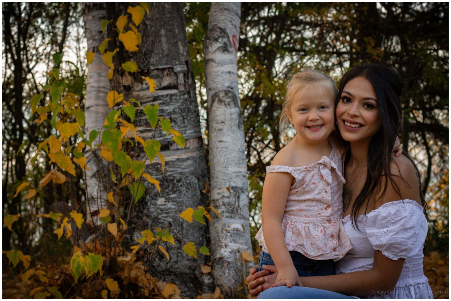 Smiling mother and toddler daughter posing together near birch trees in fall foliage.