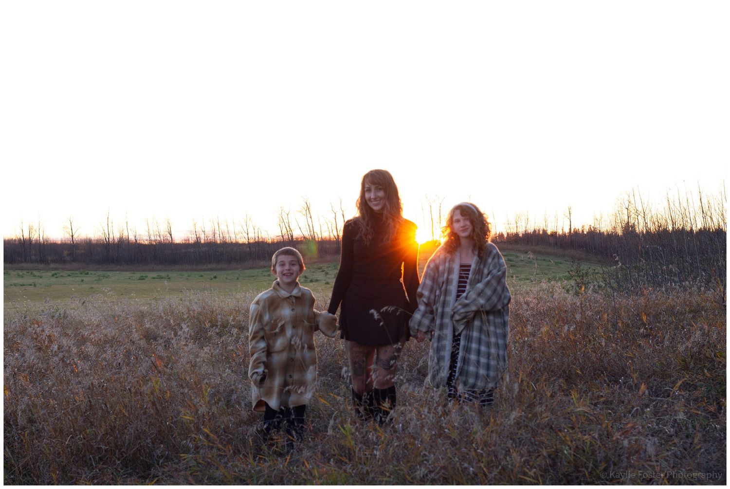 A woman and two children stand in a golden field at sunset, wearing cozy plaid coats in a serene outdoor setting.