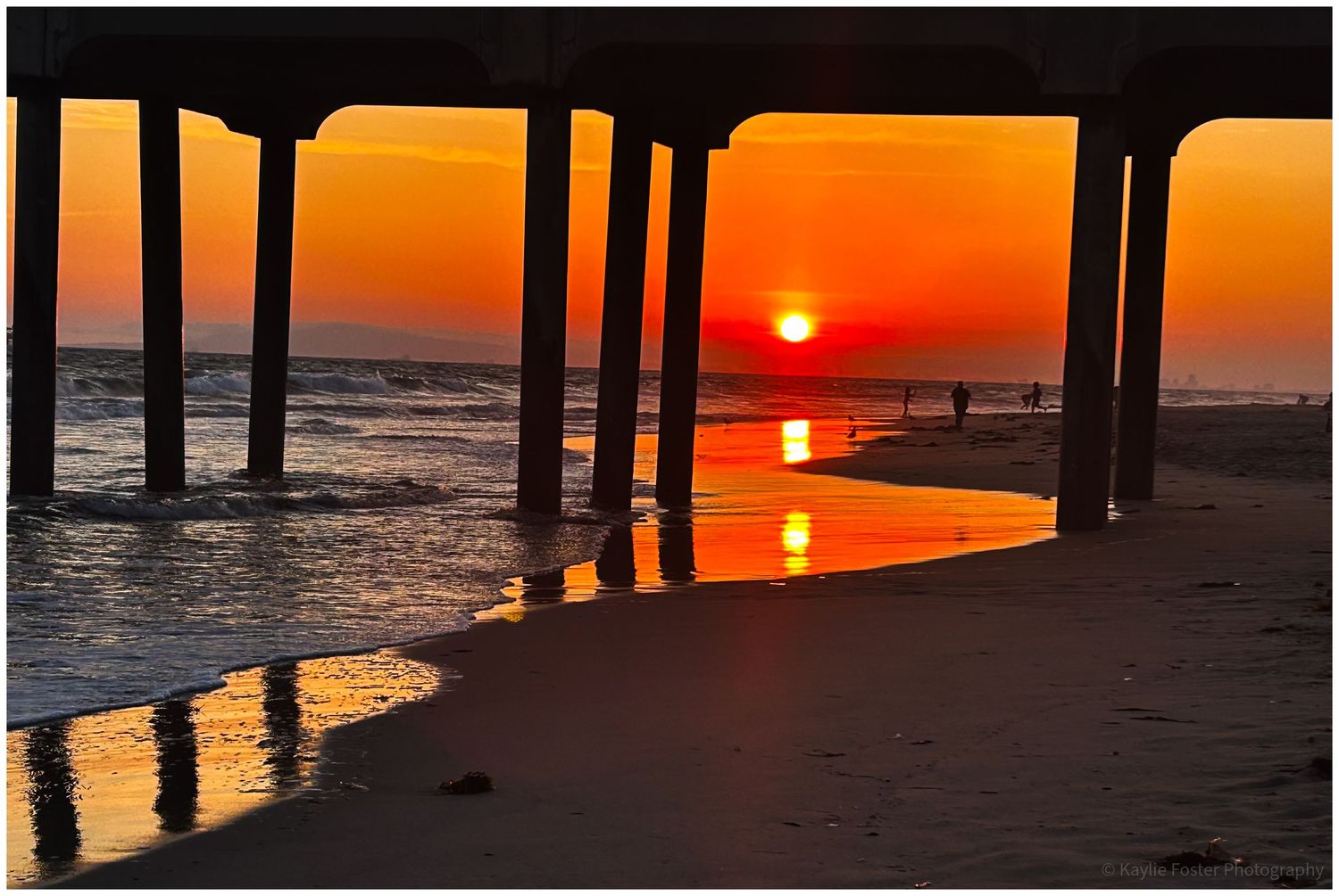 Stunning golden sunset viewed from under a beach pier with reflections on wet sand and silhouetted figures.