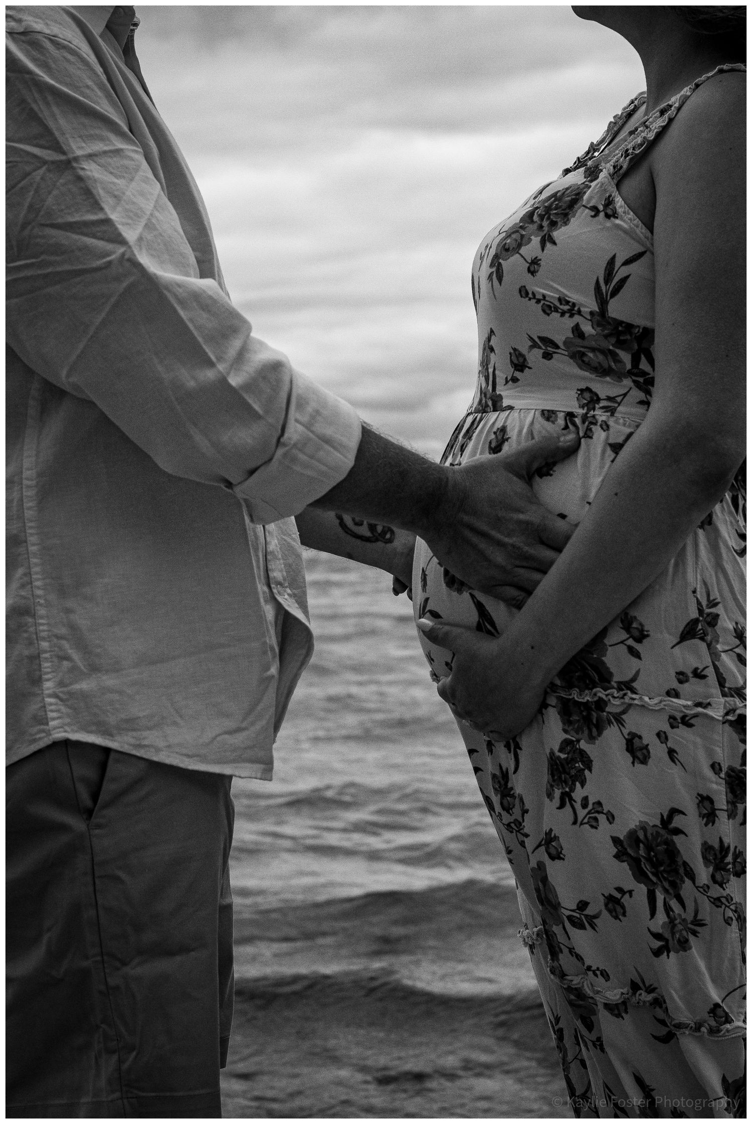 Black and white maternity photo of couple holding pregnant belly near ocean waves.