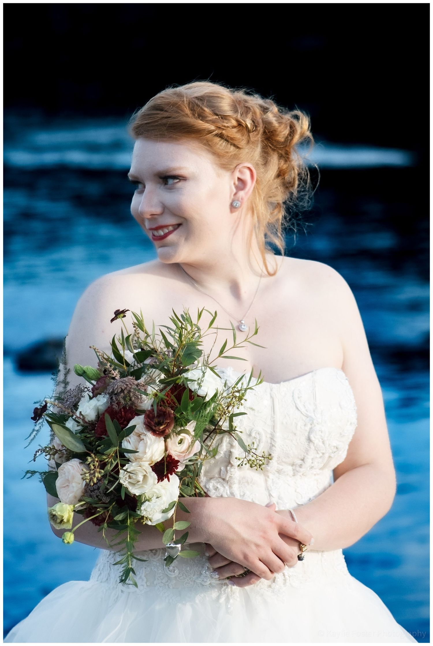 Red-haired bride in strapless lace gown holds wildflower bouquet with dark floral accents near blue ocean water.