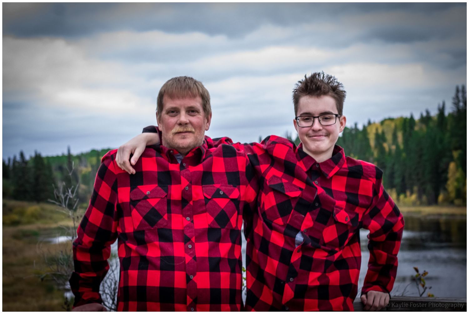 Father and son in matching red plaid shirts smiling by a river.