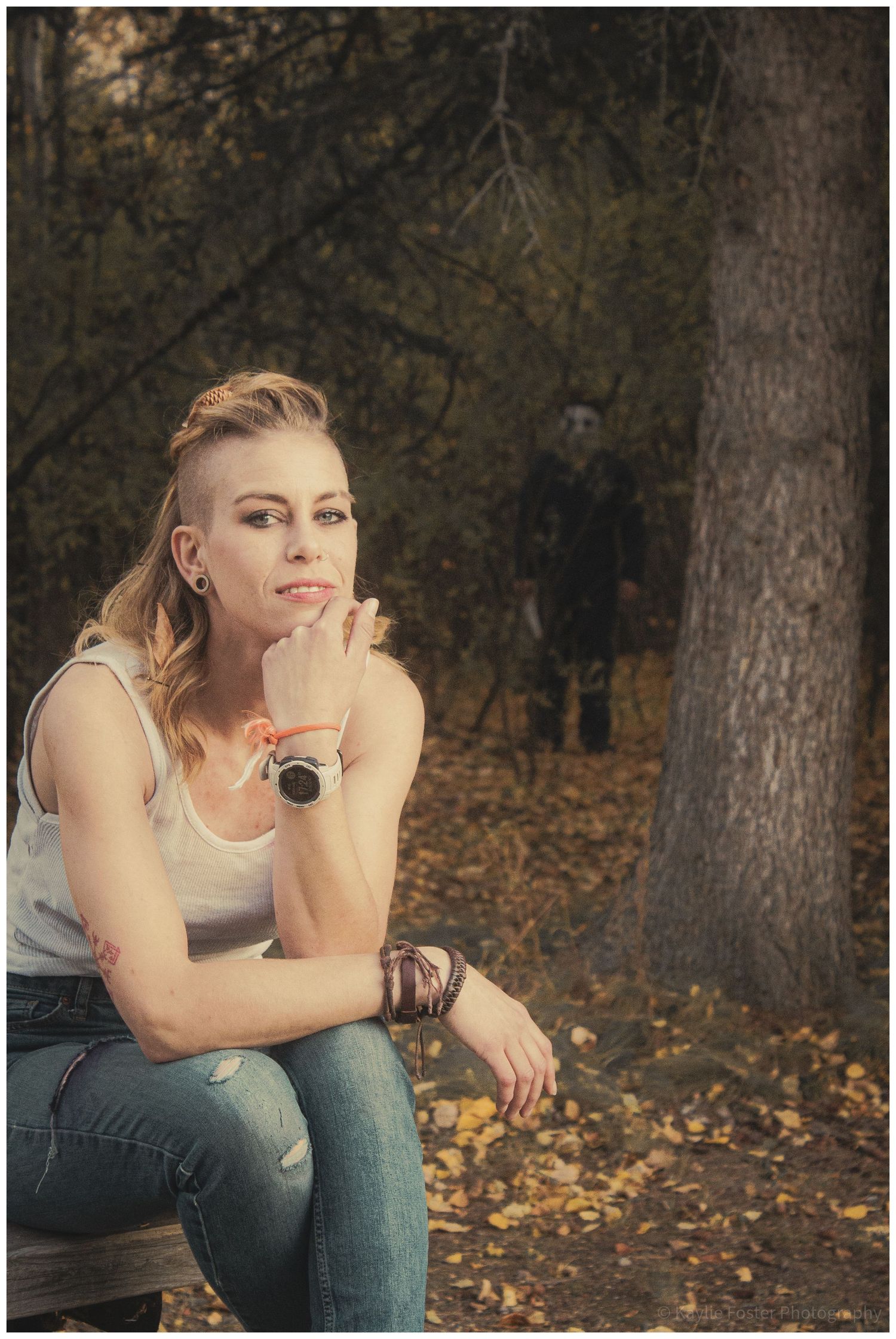 Blonde woman sitting on bench in autumn forest with mysterious masked figure lurking behind tree.