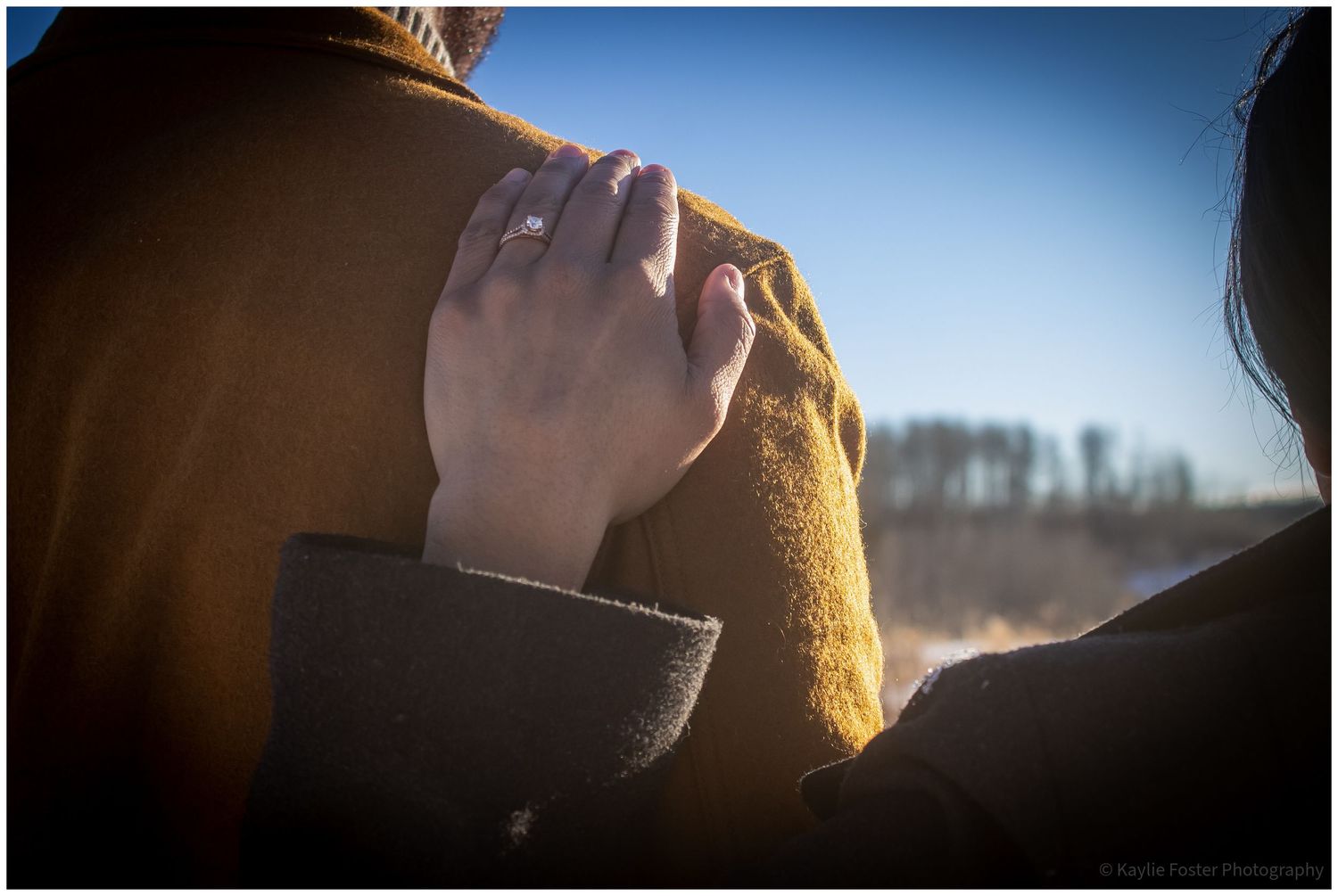 A woman's hand with an engagement ring rests on a man's shoulder outdoors in warm golden sunlight.