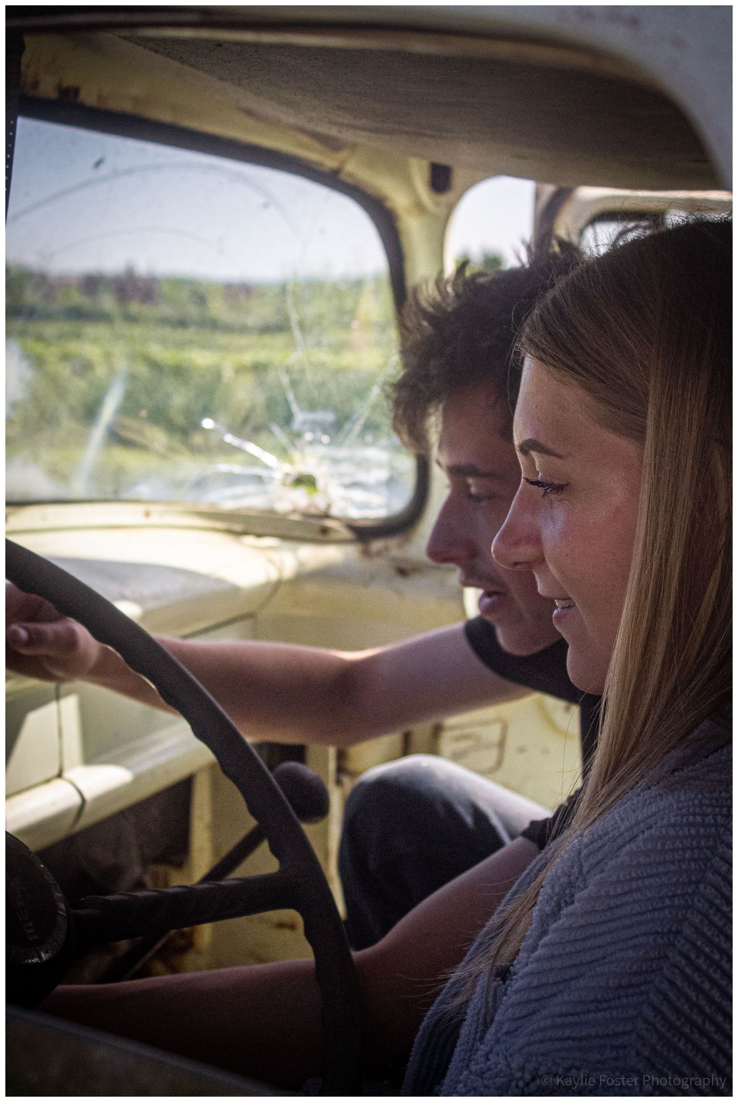 Young couple sitting inside a vintage truck with a cracked windshield, girl holding steering wheel.
