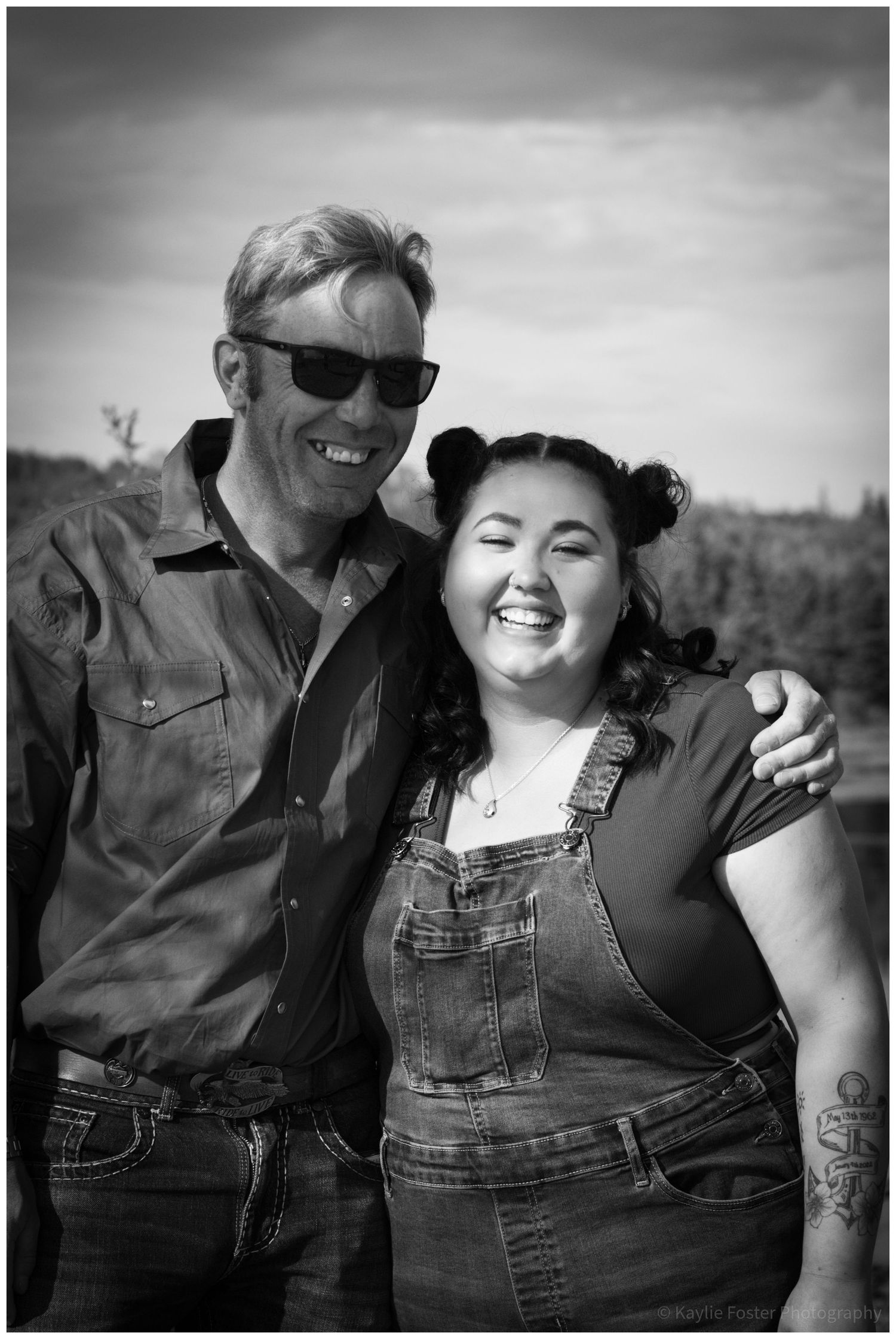 Black and white photo of a smiling man and woman posing together outdoors, woman wearing overalls with a visible arm tattoo.