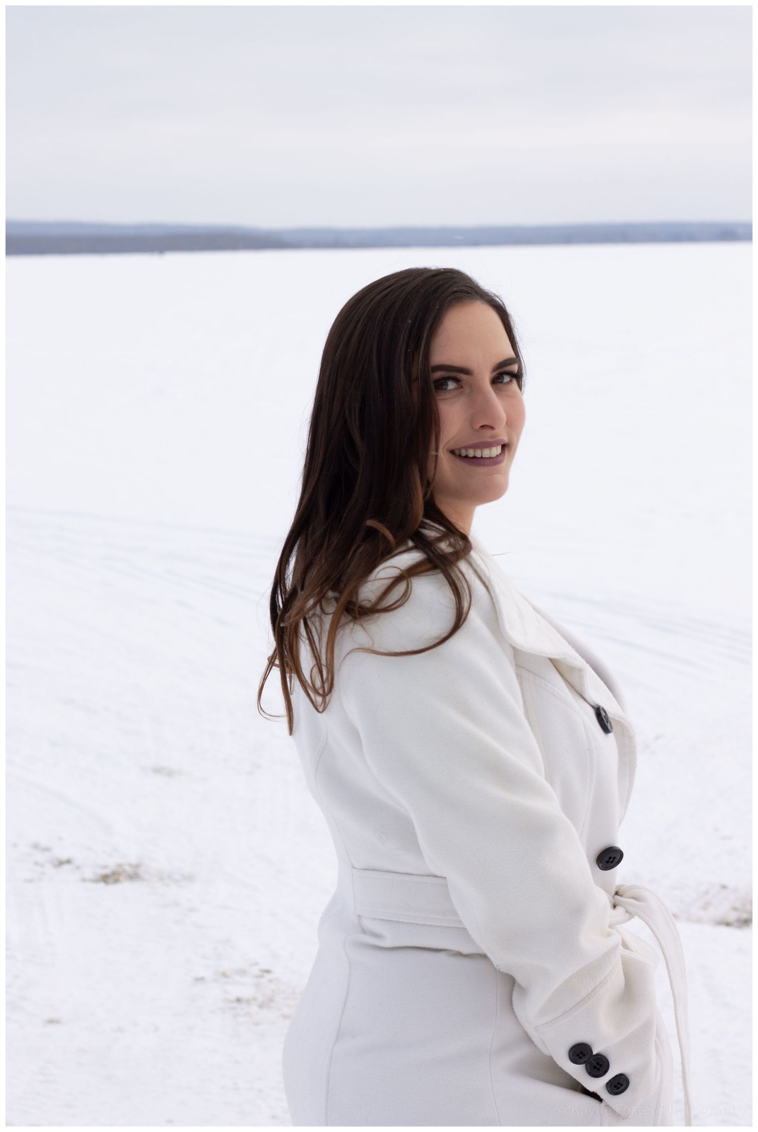 Woman in white coat smiling over shoulder in snowy winter landscape.